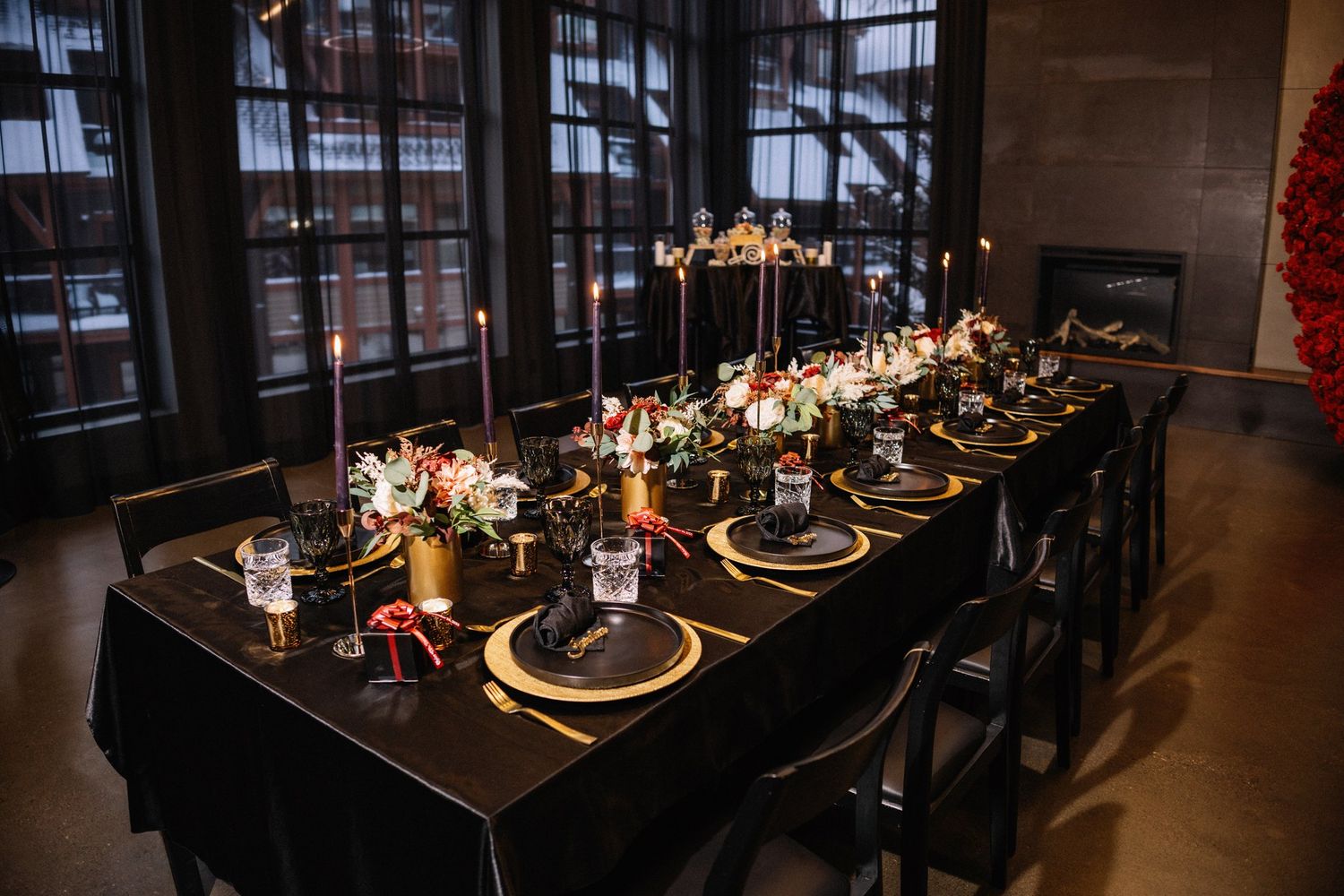 Long black reception table with gold chargers, black taper candles and red roses in front of tall windows at Mainspace