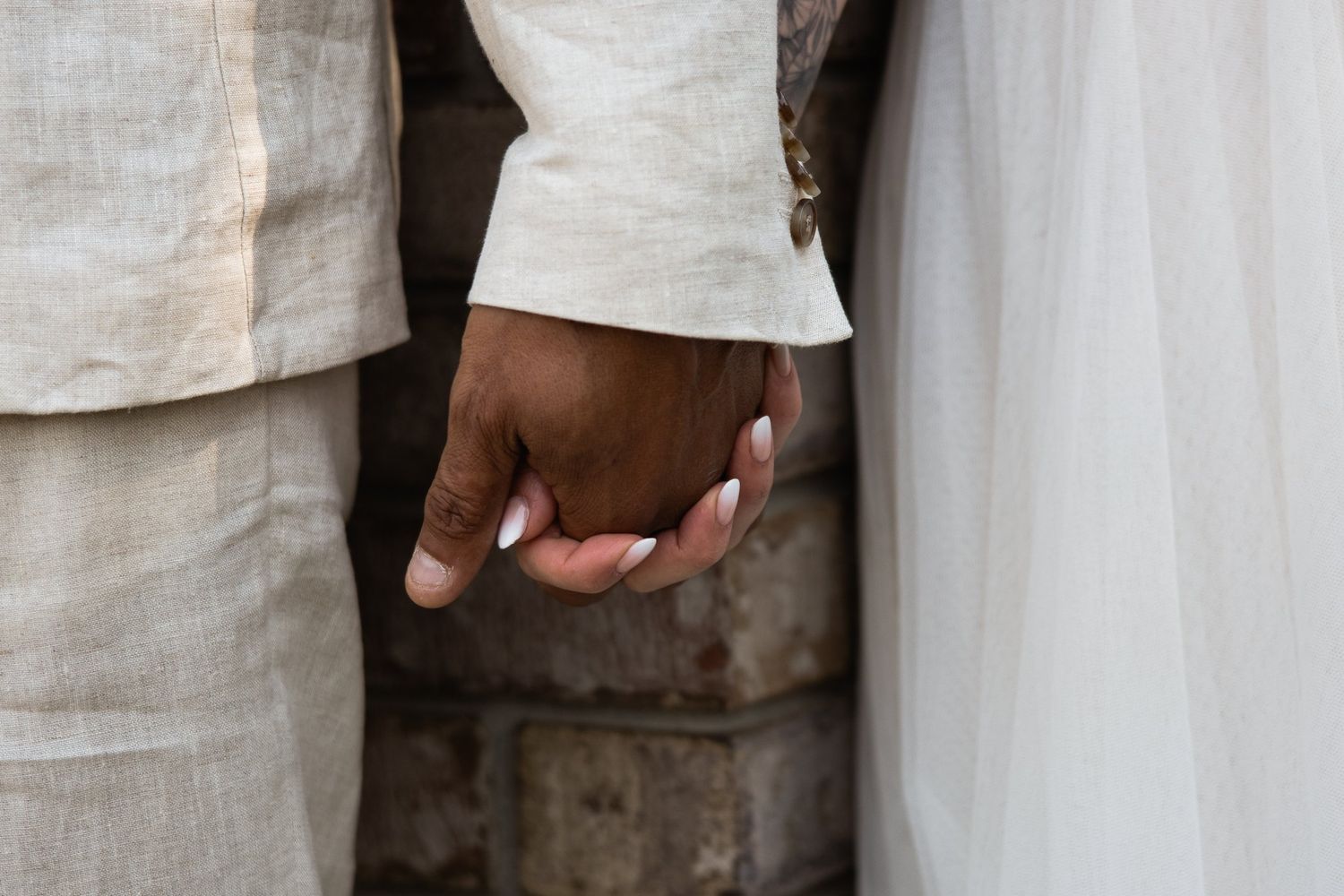 Bride's hand holding the groom's hand at his linen jacket cuff against brick, with her tulle skirt beside