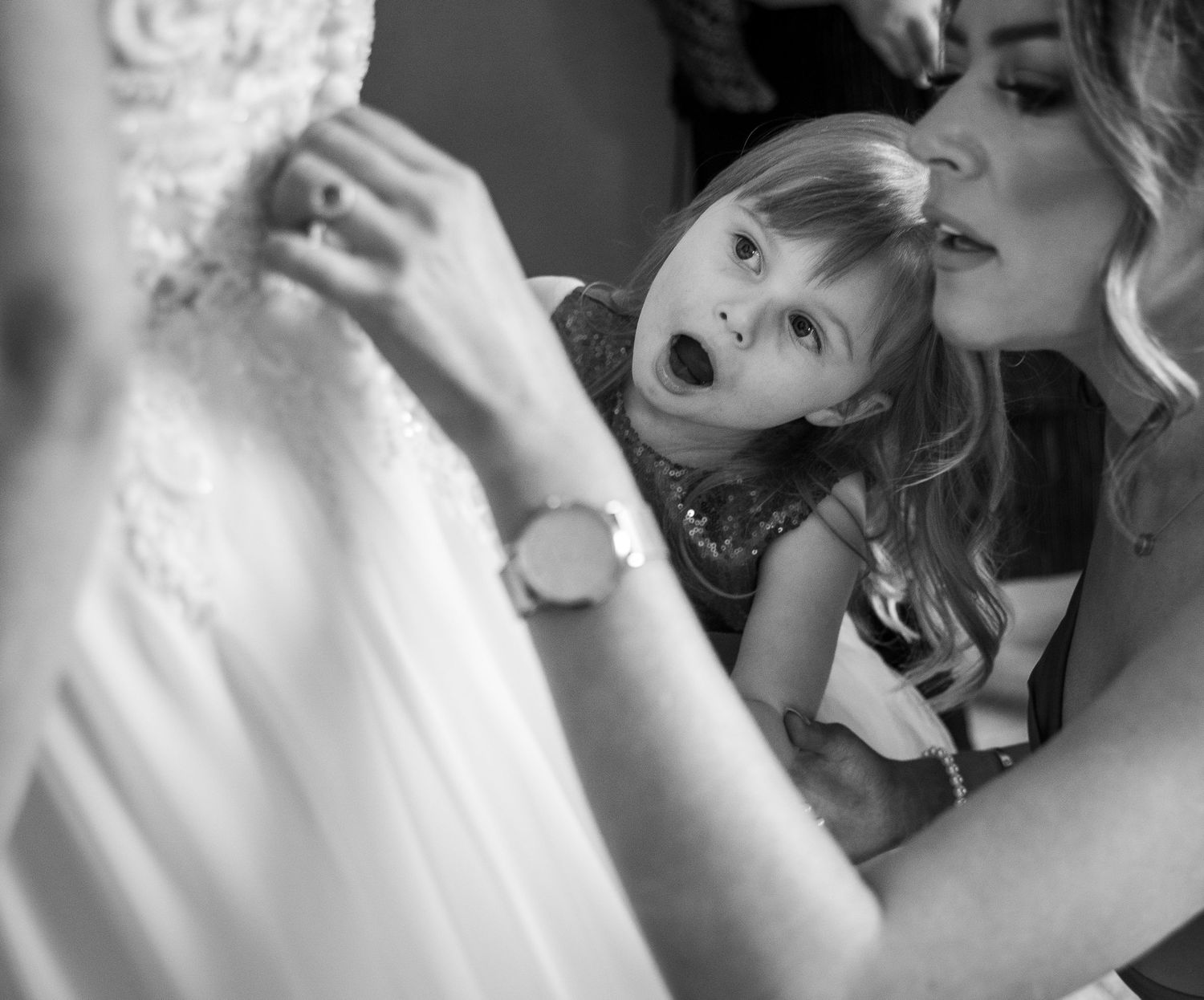 Black and white photo of a woman kissing a surprised little girl near a white wedding dress.
