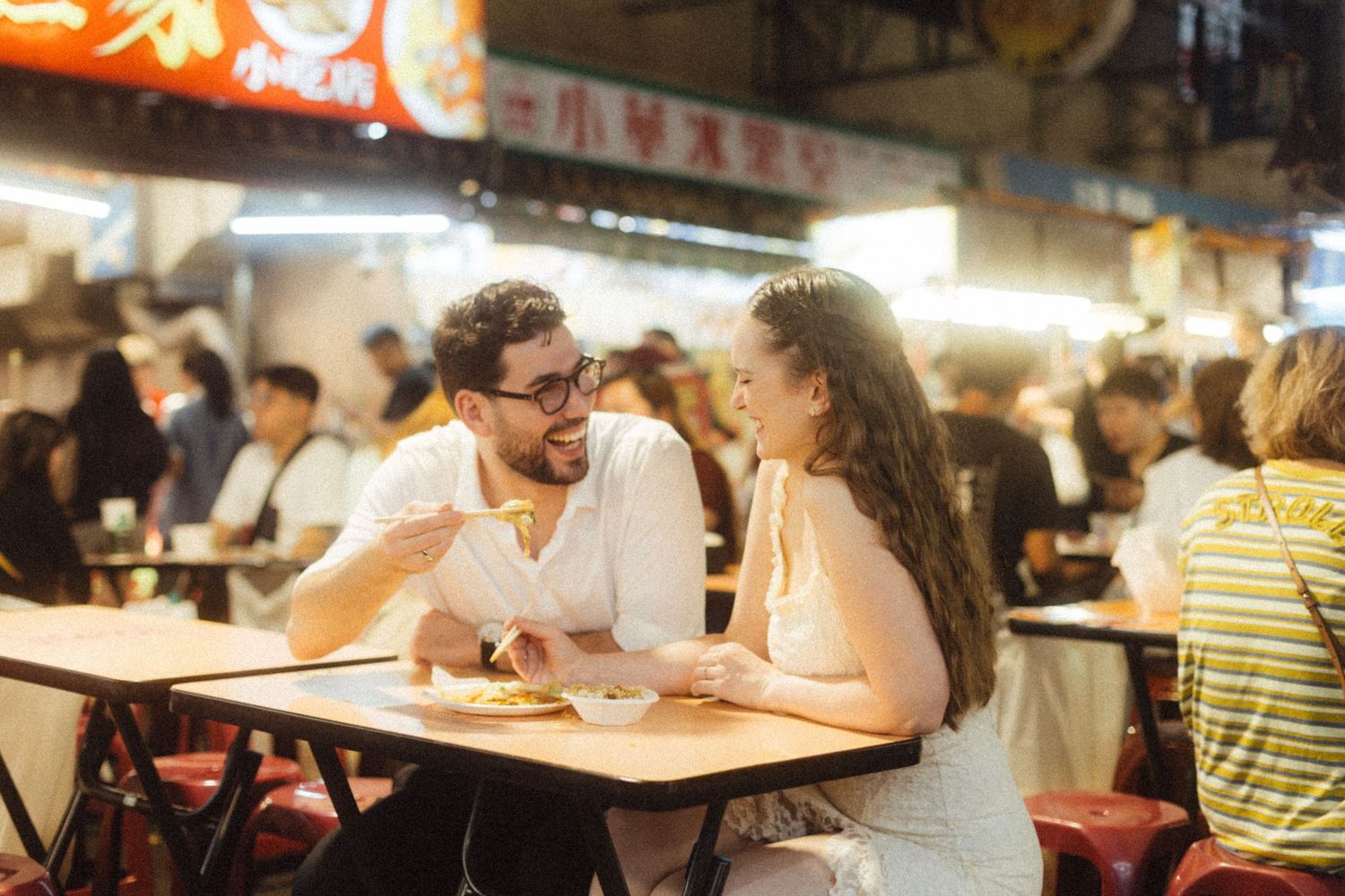 Couple enjoying street food at a busy night market in Asia, smiling and sharing a meal at an outdoor restaurant.