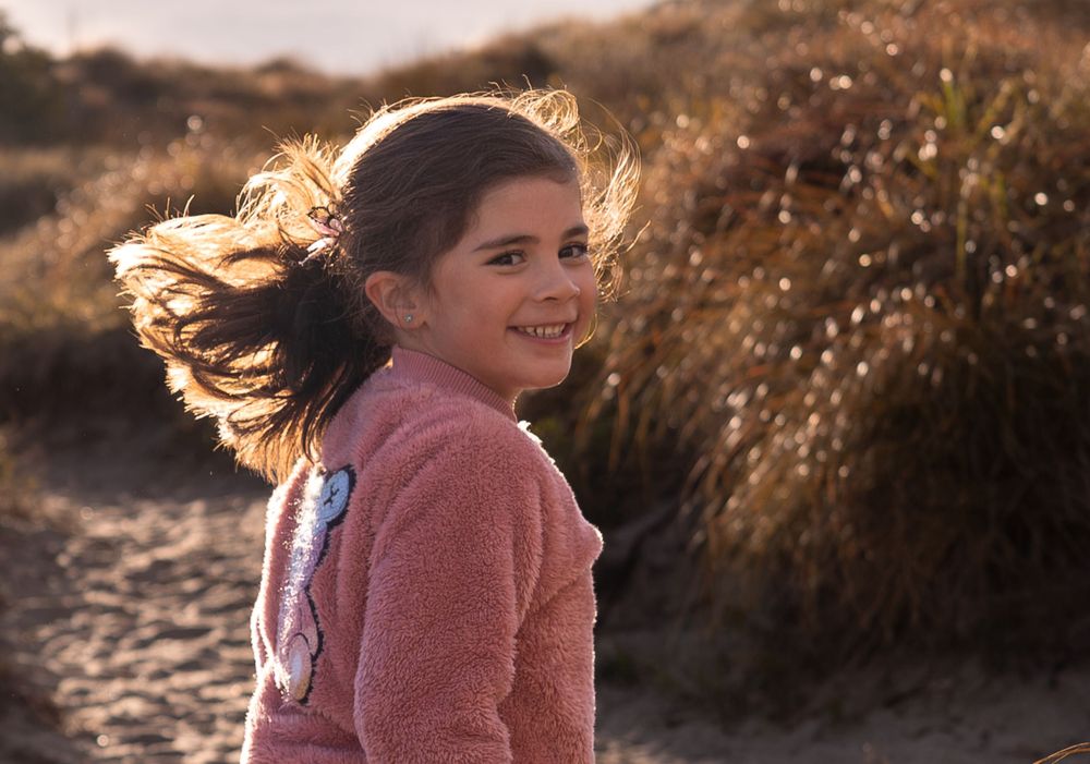Young girl smiling on a windy beach, hair blowing, wearing a pink fuzzy sweater with golden sunset light.