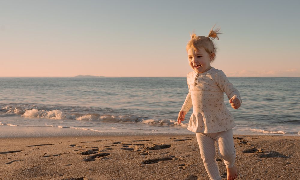 A happy toddler girl in white outfit runs along a sandy beach at golden hour with ocean waves behind her.