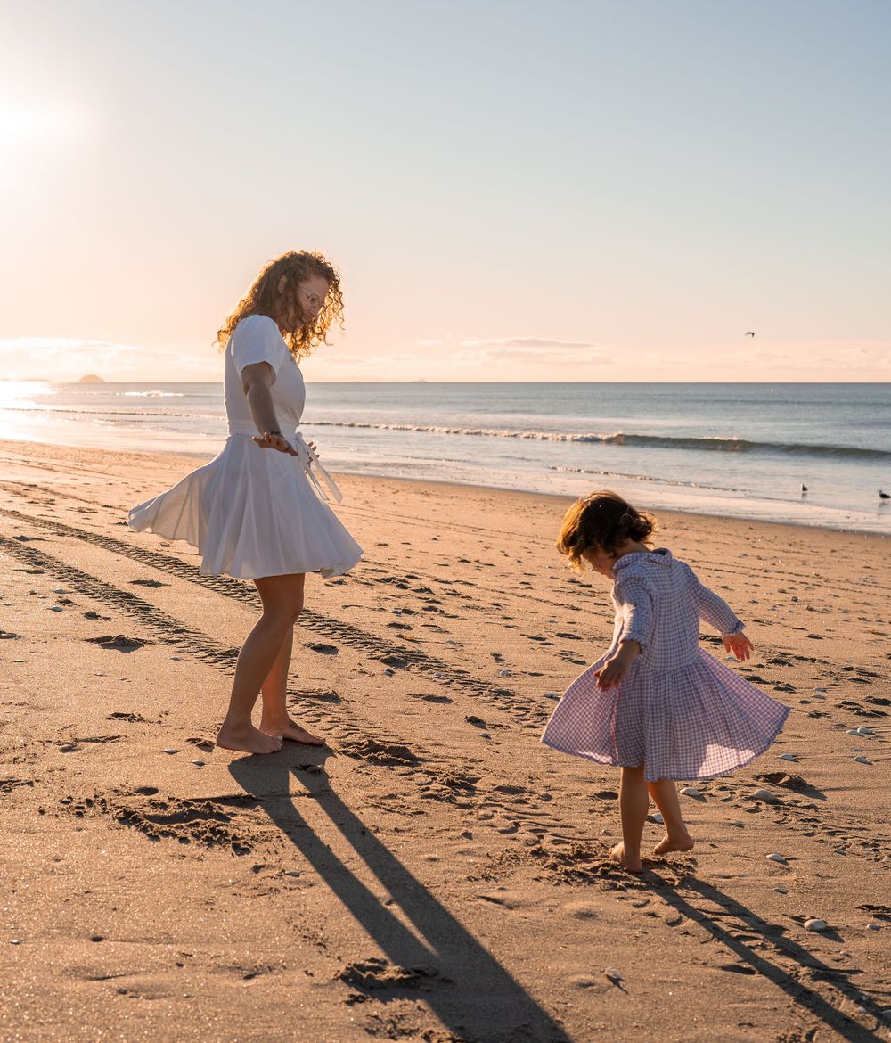 A mother and daughter in matching white dresses dance barefoot on a sandy beach at golden sunset.