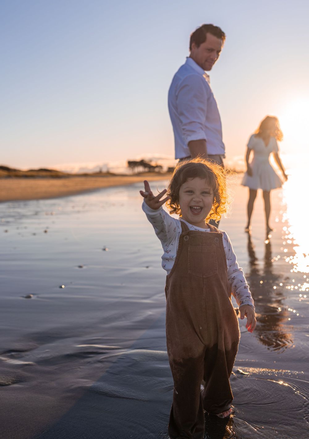 A joyful toddler in brown overalls plays on a sunlit beach at golden hour with family in the background.