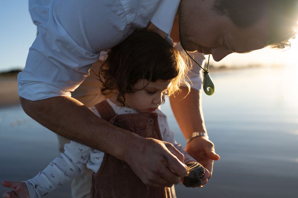 Father and toddler examining a small rock together on a sunlit beach at dusk.