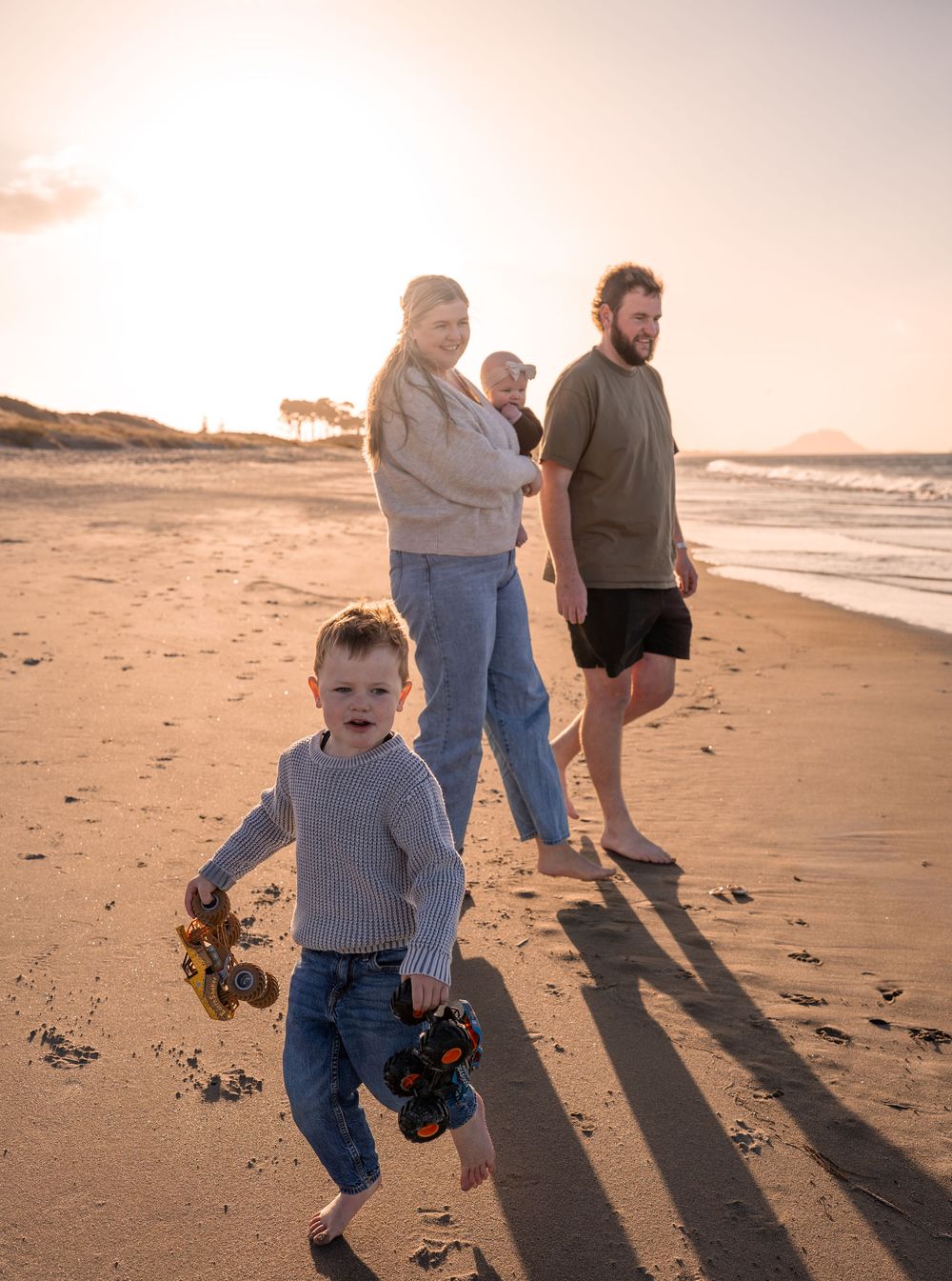 Family walking on a sandy beach at sunset, young boy holding toy trucks in foreground, parents with baby strolling behind.