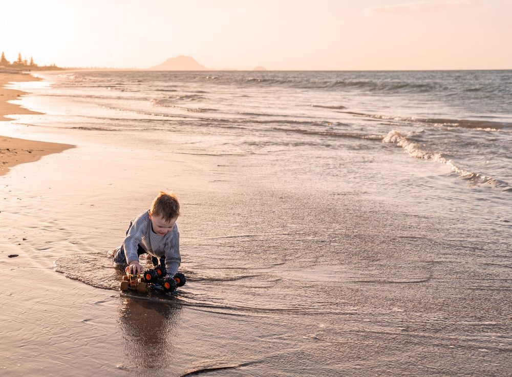 A young child plays alone on a sandy beach at sunset, crouching near the shoreline with gentle waves nearby.