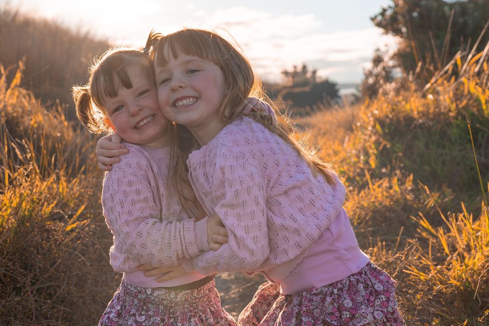 Two young girls hugging and smiling in a golden sunlit field at sunset, wearing pink floral outfits.