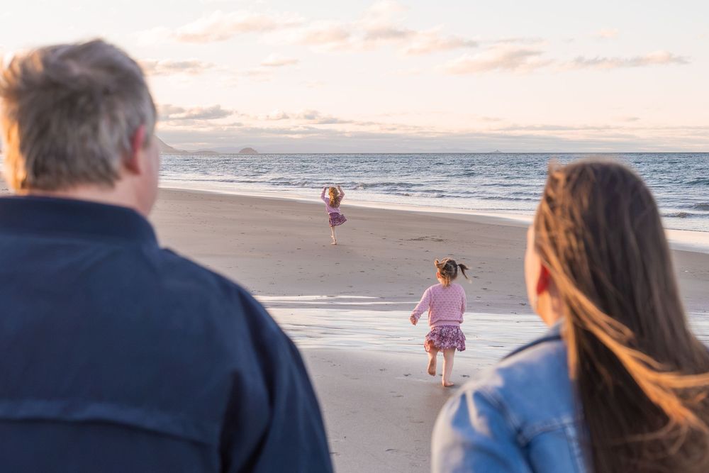 Two adults watch children playing on a sandy beach at sunset with gentle ocean waves in the background.