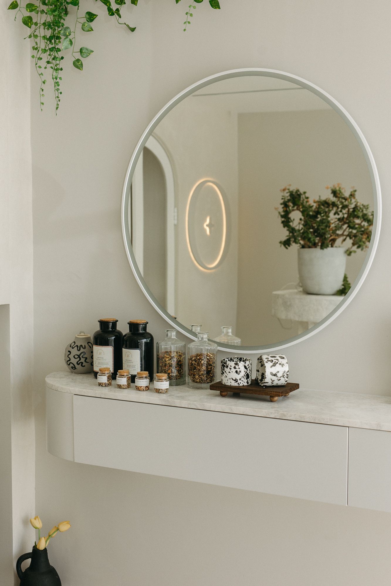 Minimalist white floating shelf with round mirror, dark glass bottles, and small jars in a bright, modern bathroom setting.
