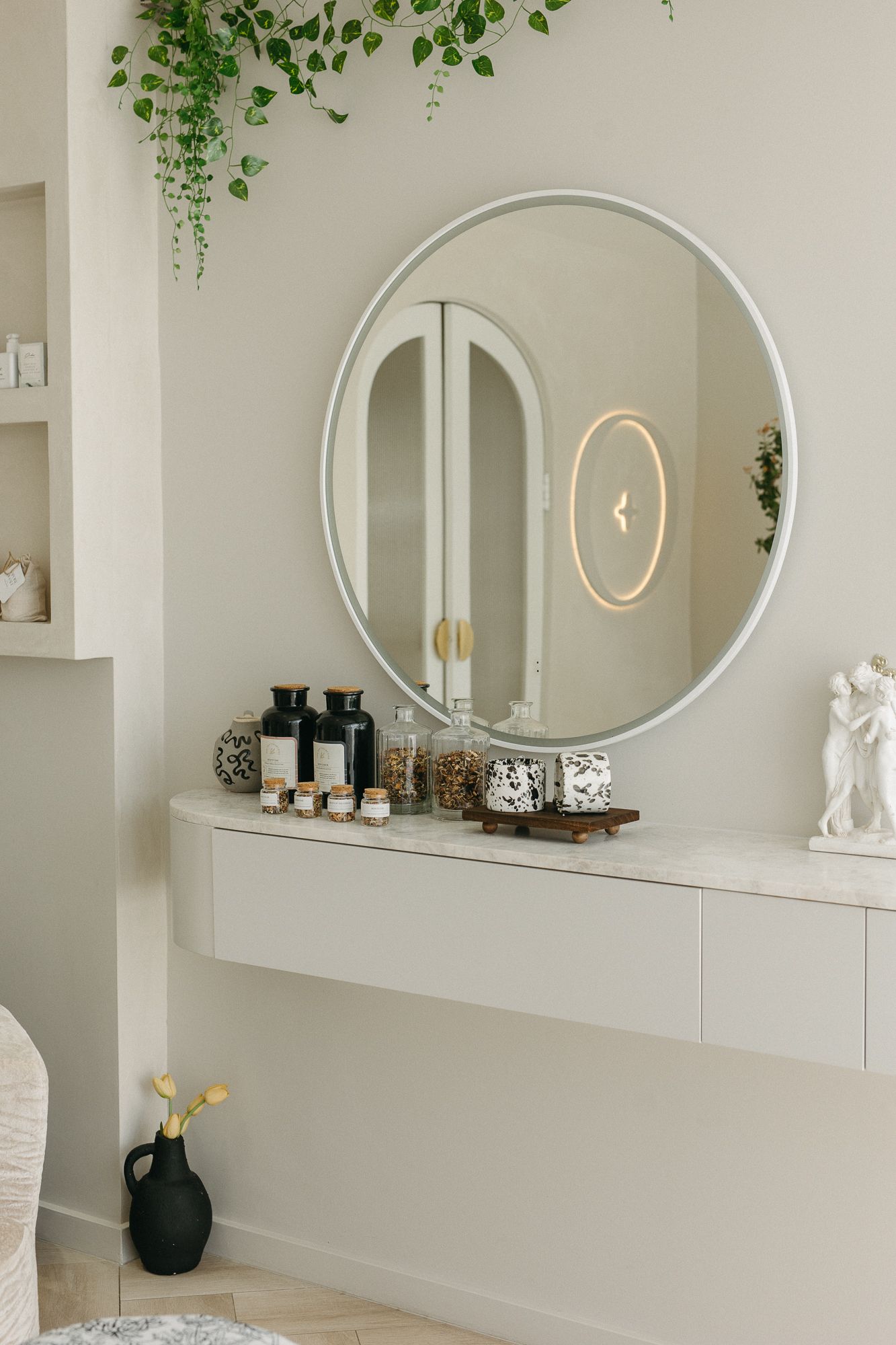 Elegant vanity area with round mirror, beauty products, and trailing green plants in a neutral spa-like room.