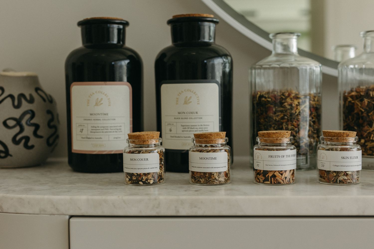 Assorted herbal tea jars with cork lids and white labels arranged on a marble countertop next to dark glass bottles.