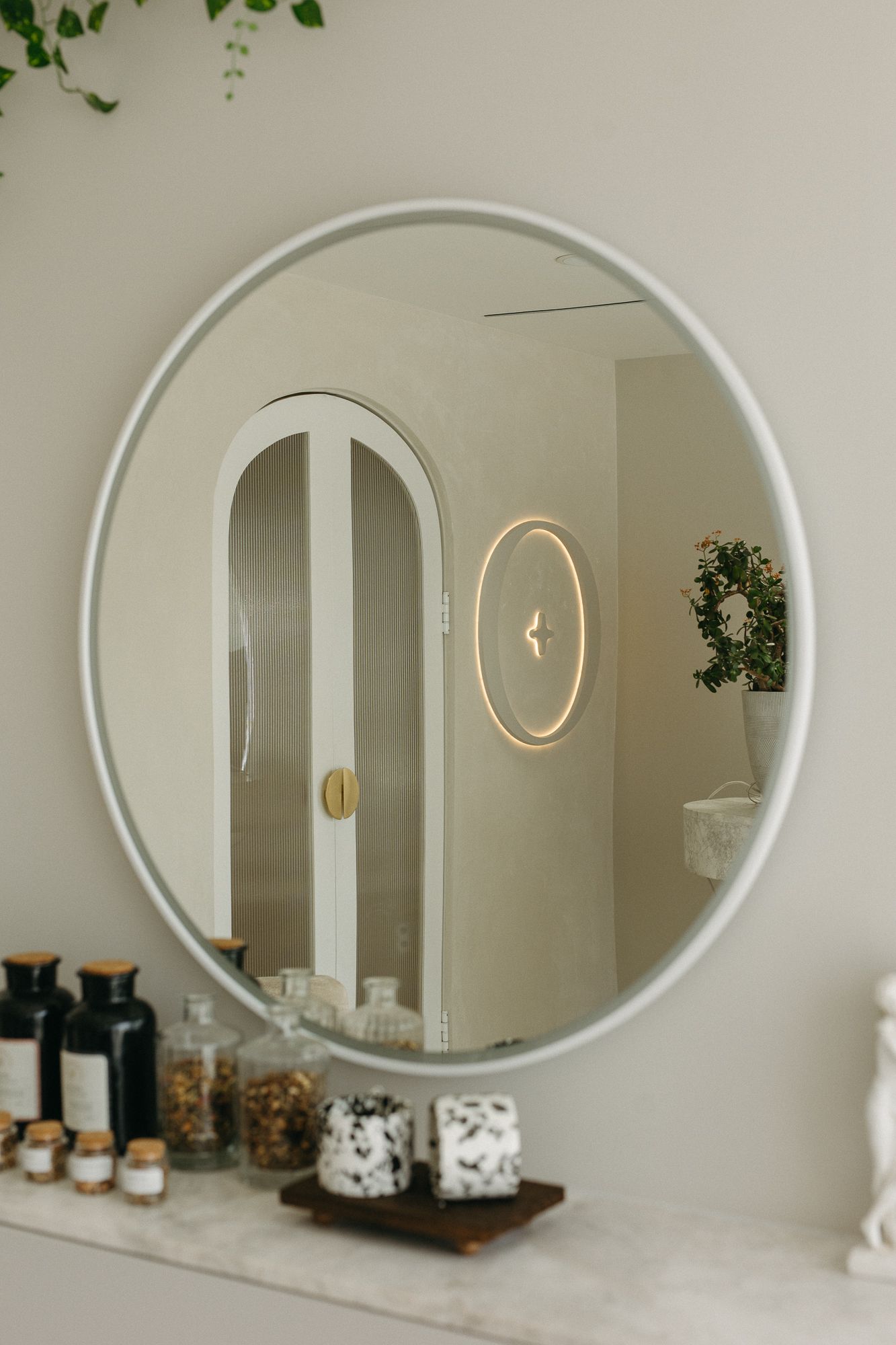 Round mirror reflecting arched white door and glowing circular light, above marble shelf with decorative glass jars.