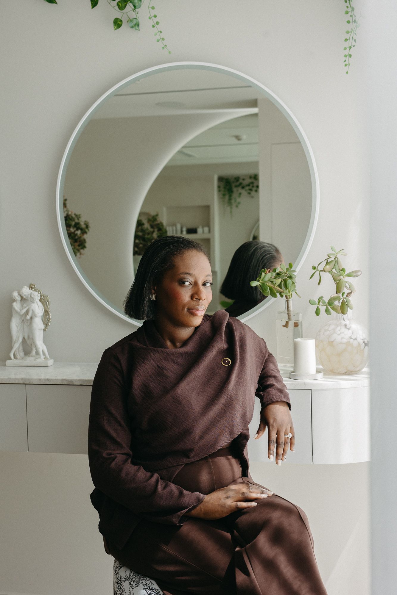 A young Black woman in a brown outfit sits thoughtfully before a round mirror with plants and decor in a bright room.