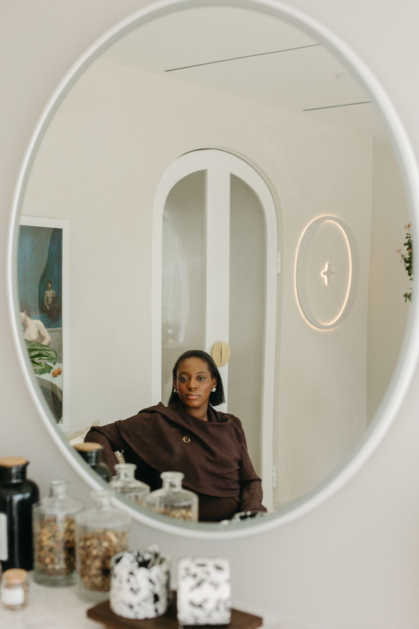 Woman in brown jacket reflected in oval mirror above decorative jars in a modern white interior space.