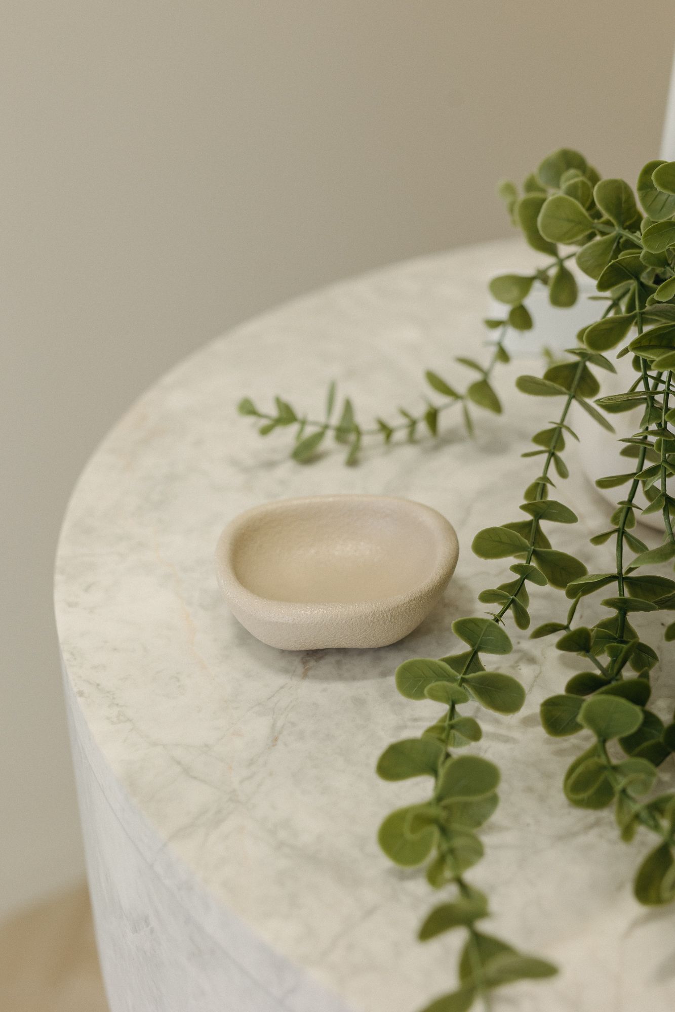 Small beige ceramic dish on a round white marble table with trailing green eucalyptus plant.