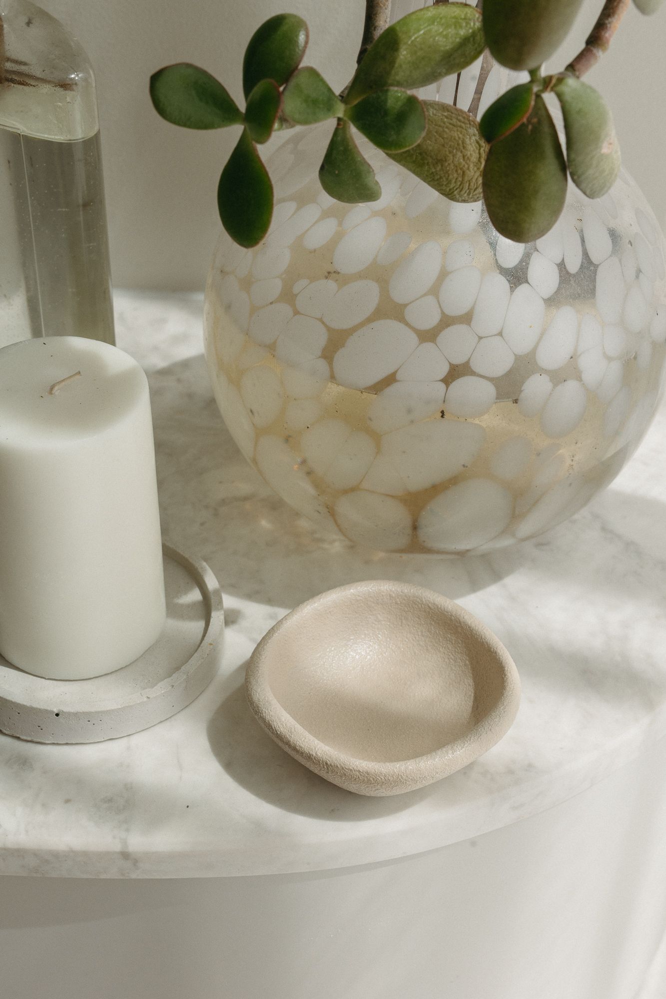 Small beige stone dish on marble surface beside white pillar candle and mosaic vase with green succulent branches.