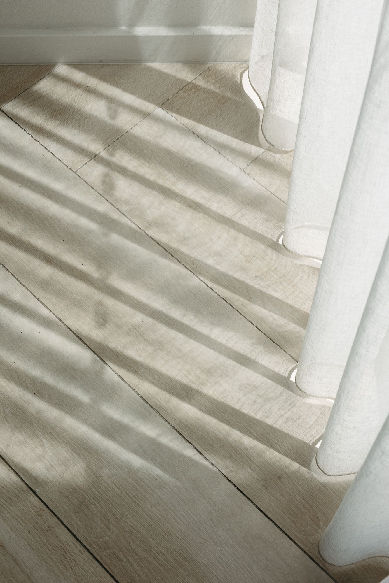 Sheer white curtains casting striped shadows on light wood floor in bright natural sunlight.