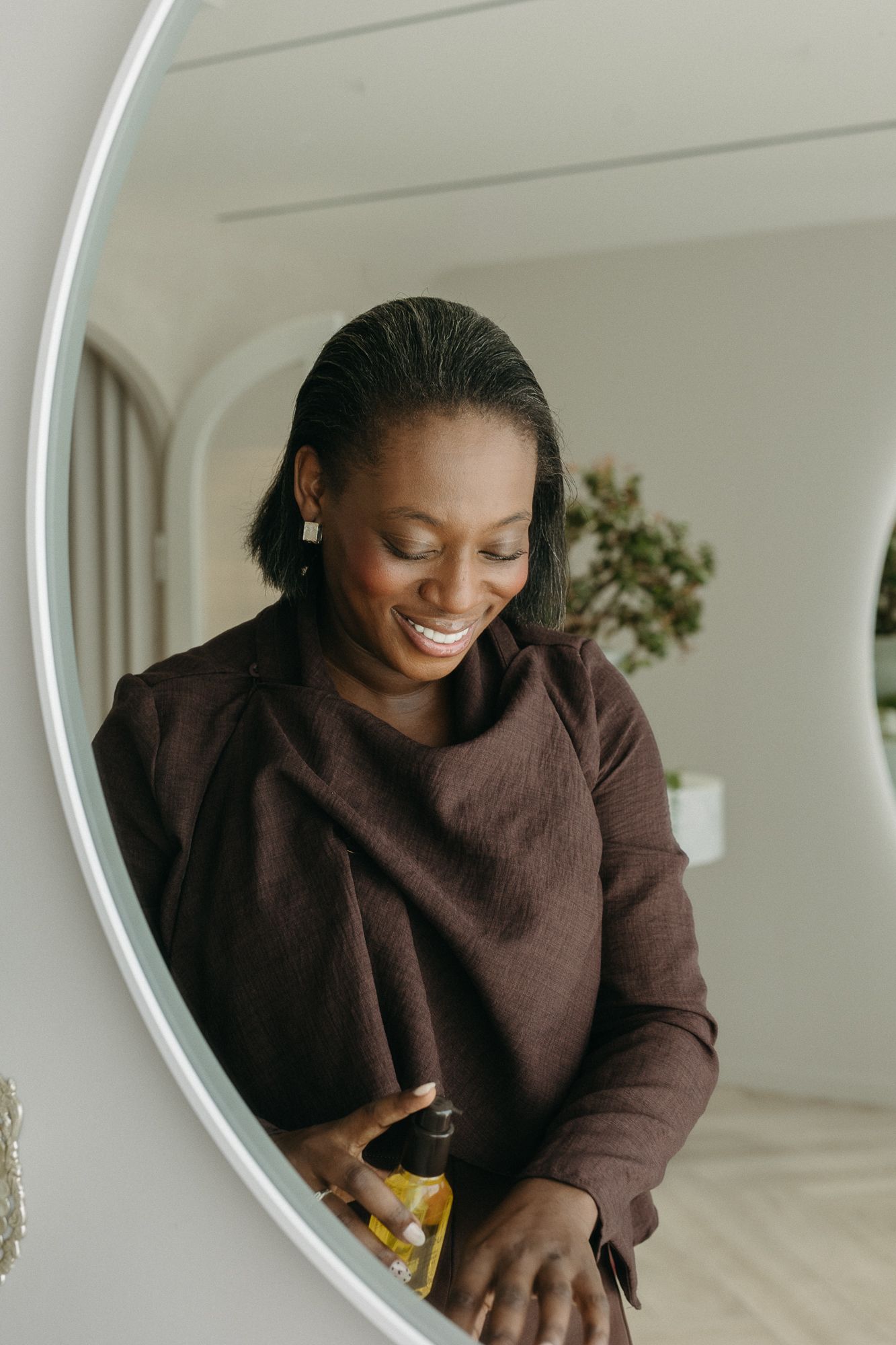 Smiling Black woman in brown sweater holding a beauty oil product while looking down in a bright bathroom mirror.