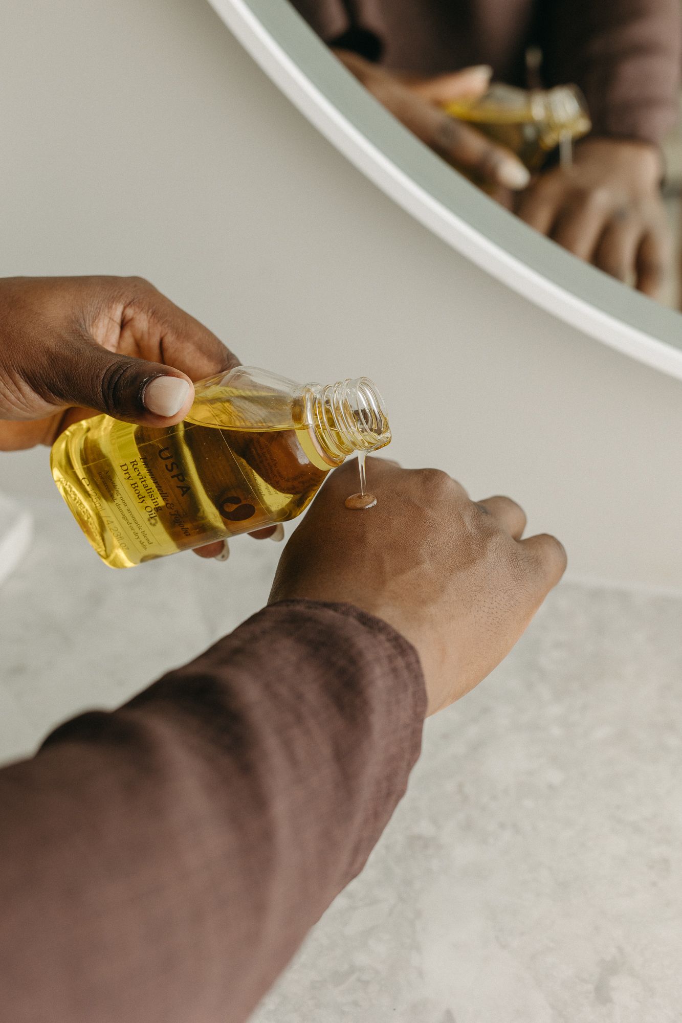 Person pouring golden oil from a small bottle into their hand, reflected in a round mirror above a marble surface.