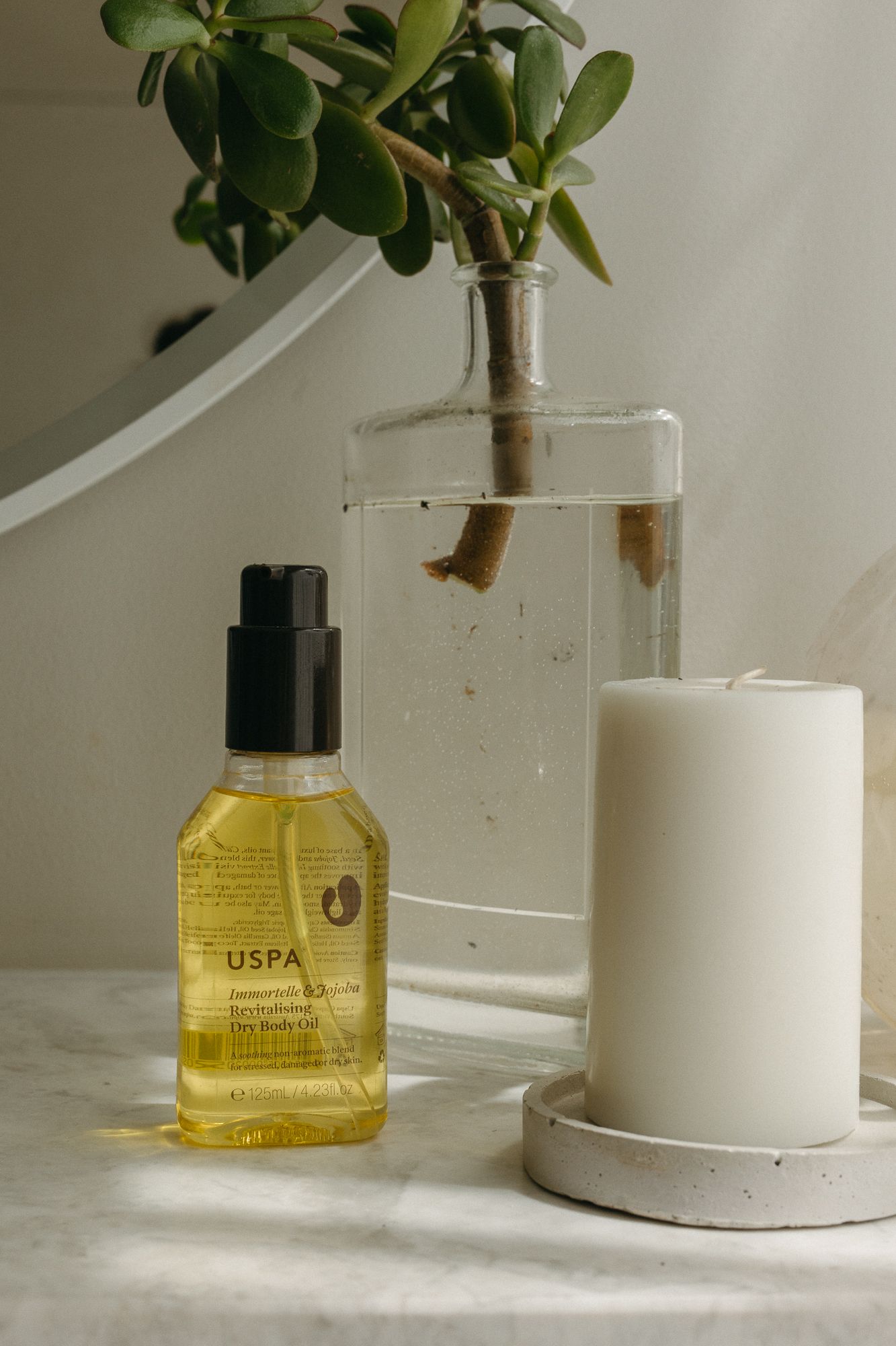 USPA body oil in yellow bottle beside white pillar candle and plant in glass vase on marble surface.