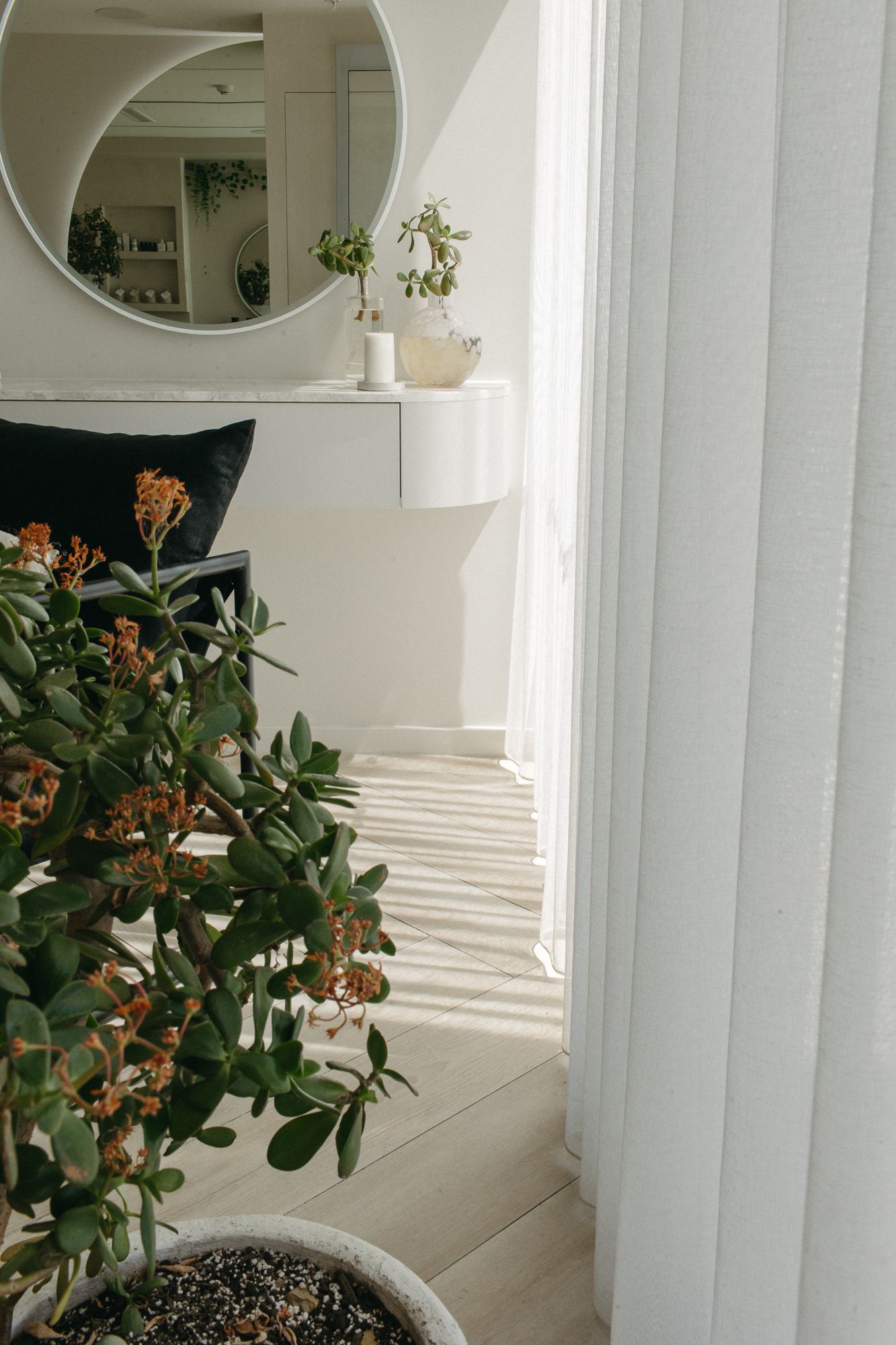 Bright Scandinavian living room with a potted jade plant, white curtains, round mirror, and minimalist white console table.