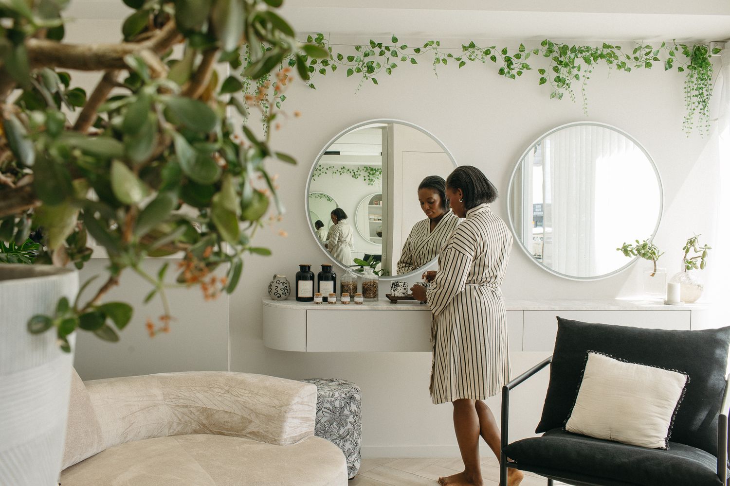 Woman in robe arranging beauty products on vanity table with round mirrors and lush green plants in bright white room.