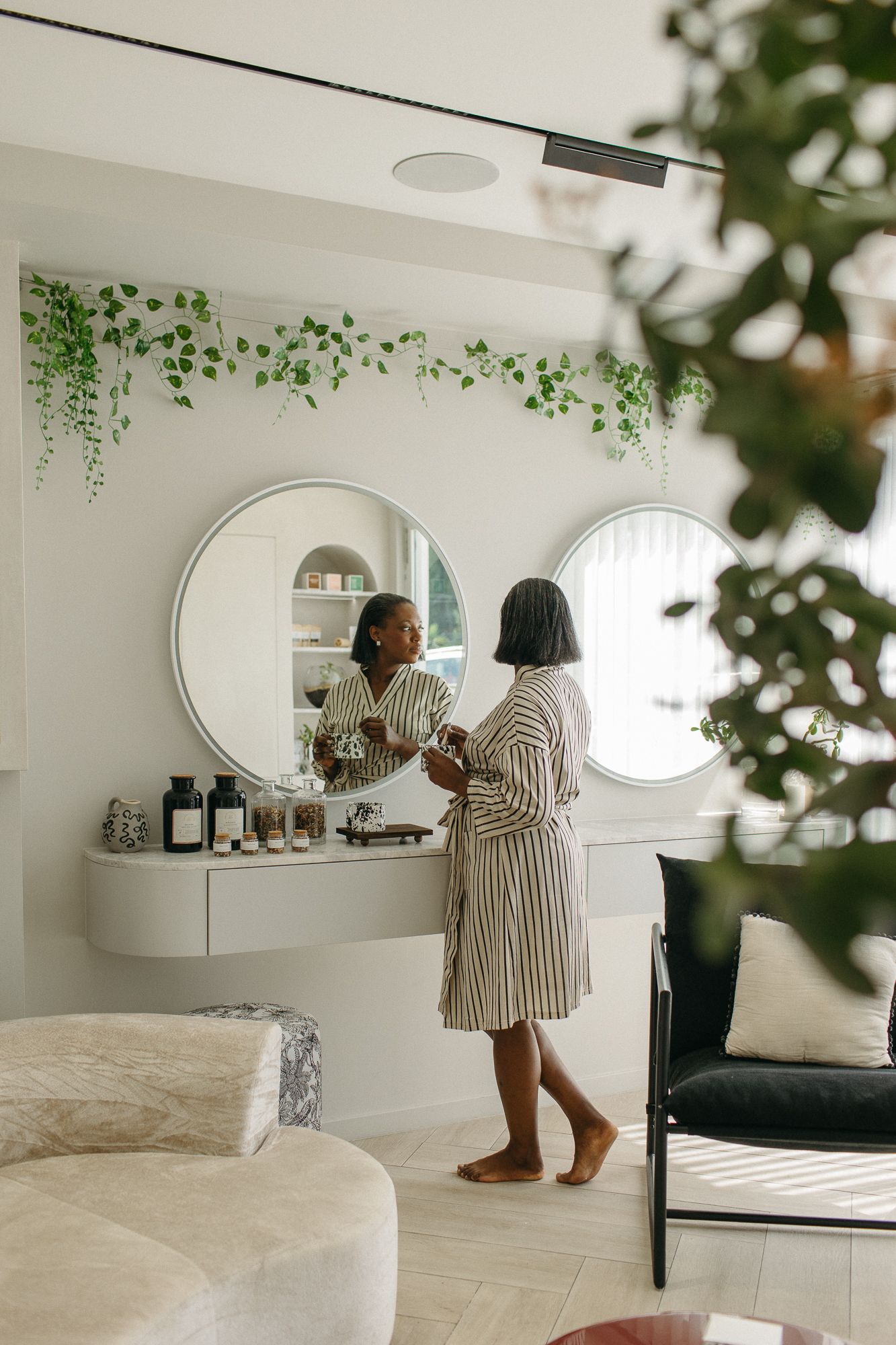 Woman in striped robe standing at vanity mirror in elegant spa-like bathroom with green trailing plants.