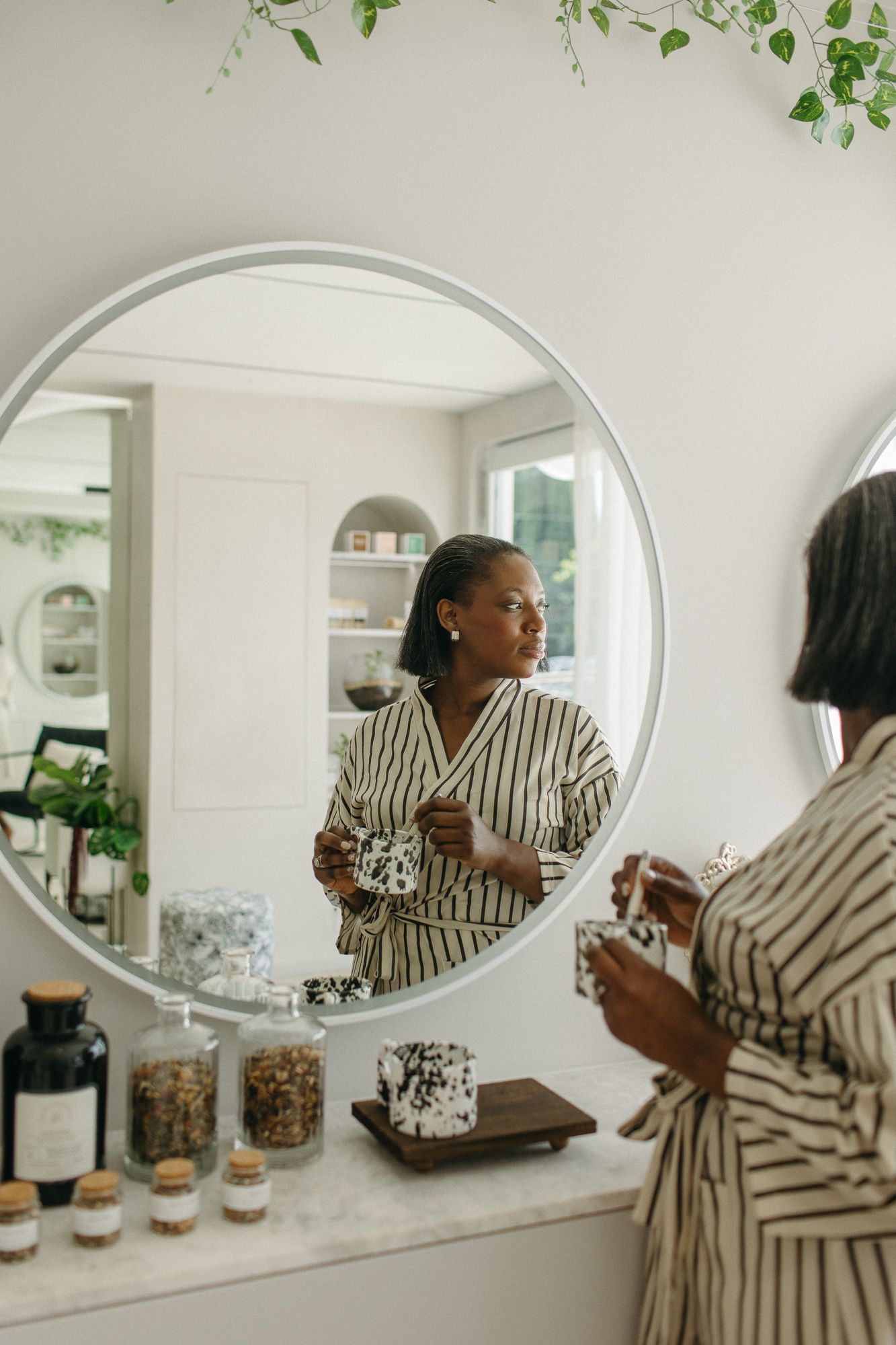 A woman in a striped robe holds a candle while looking at her reflection in a round mirror above a marble vanity.
