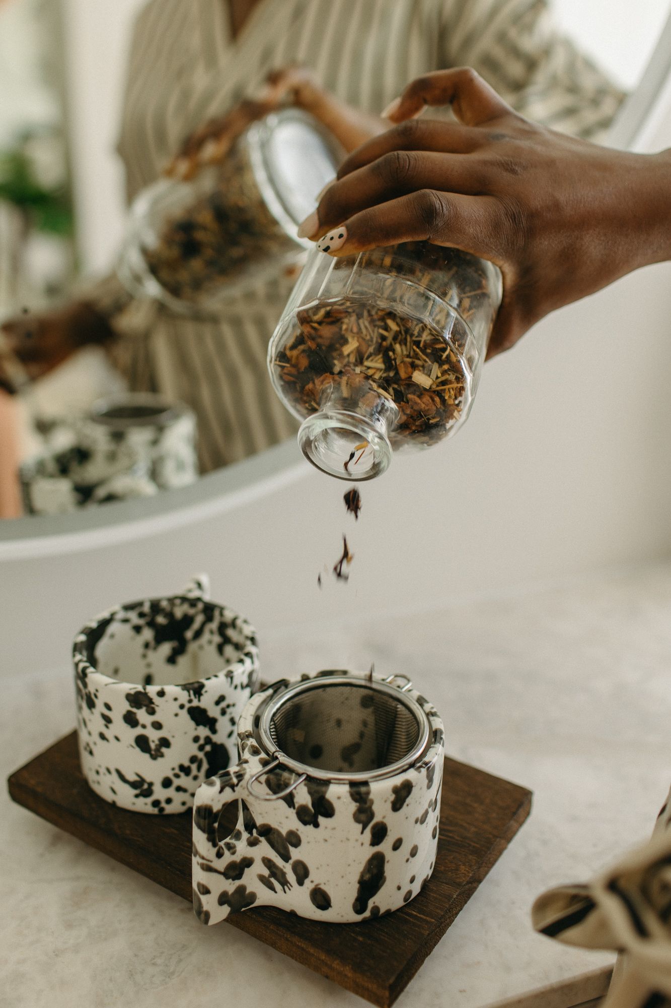 A hand pours loose leaf tea from a glass jar into a speckled ceramic teapot with a metal infuser on a wooden board.