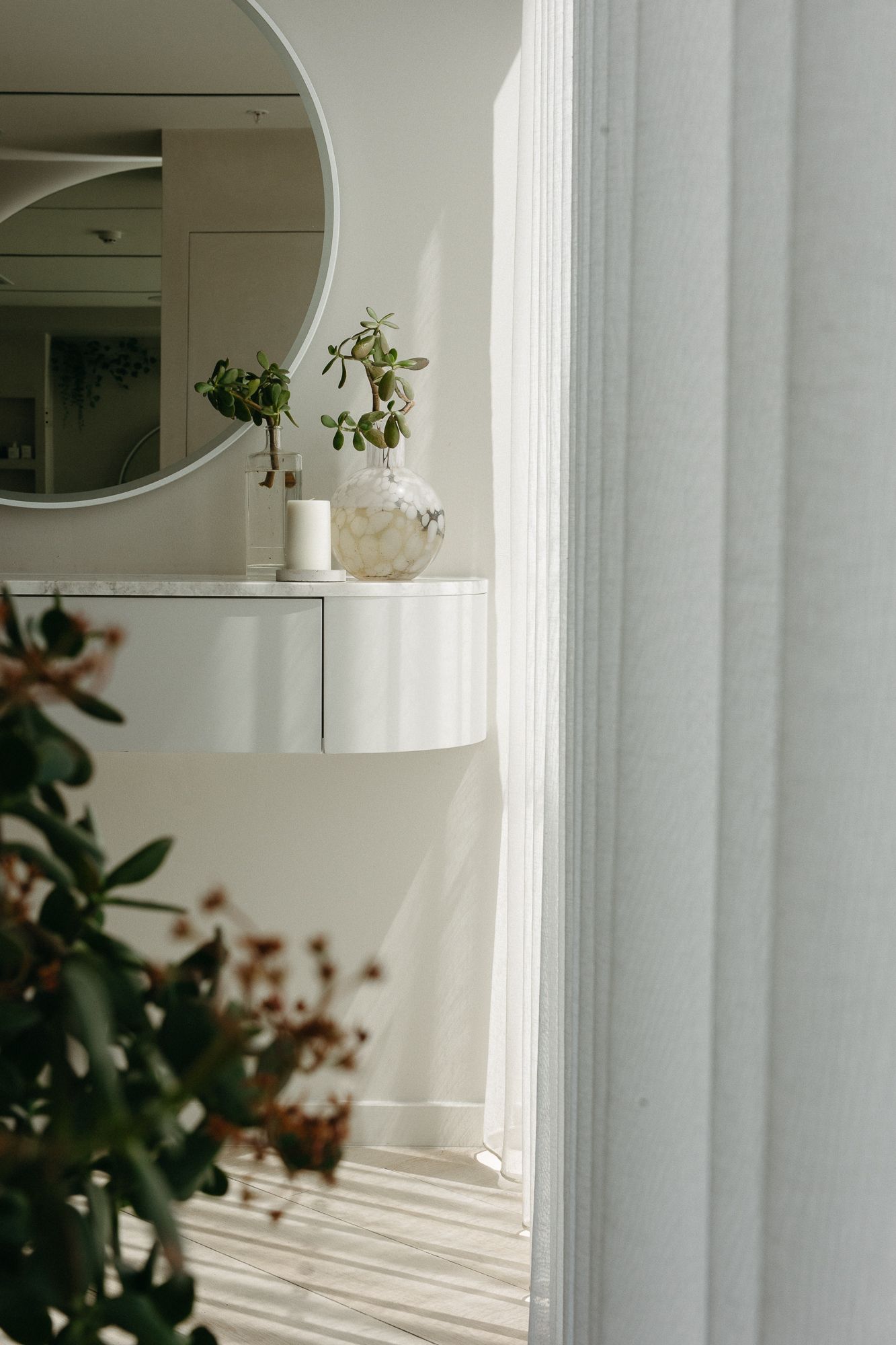 Minimalist white entryway with round mirror, floating console, glass vase with green plant, and soft natural light.