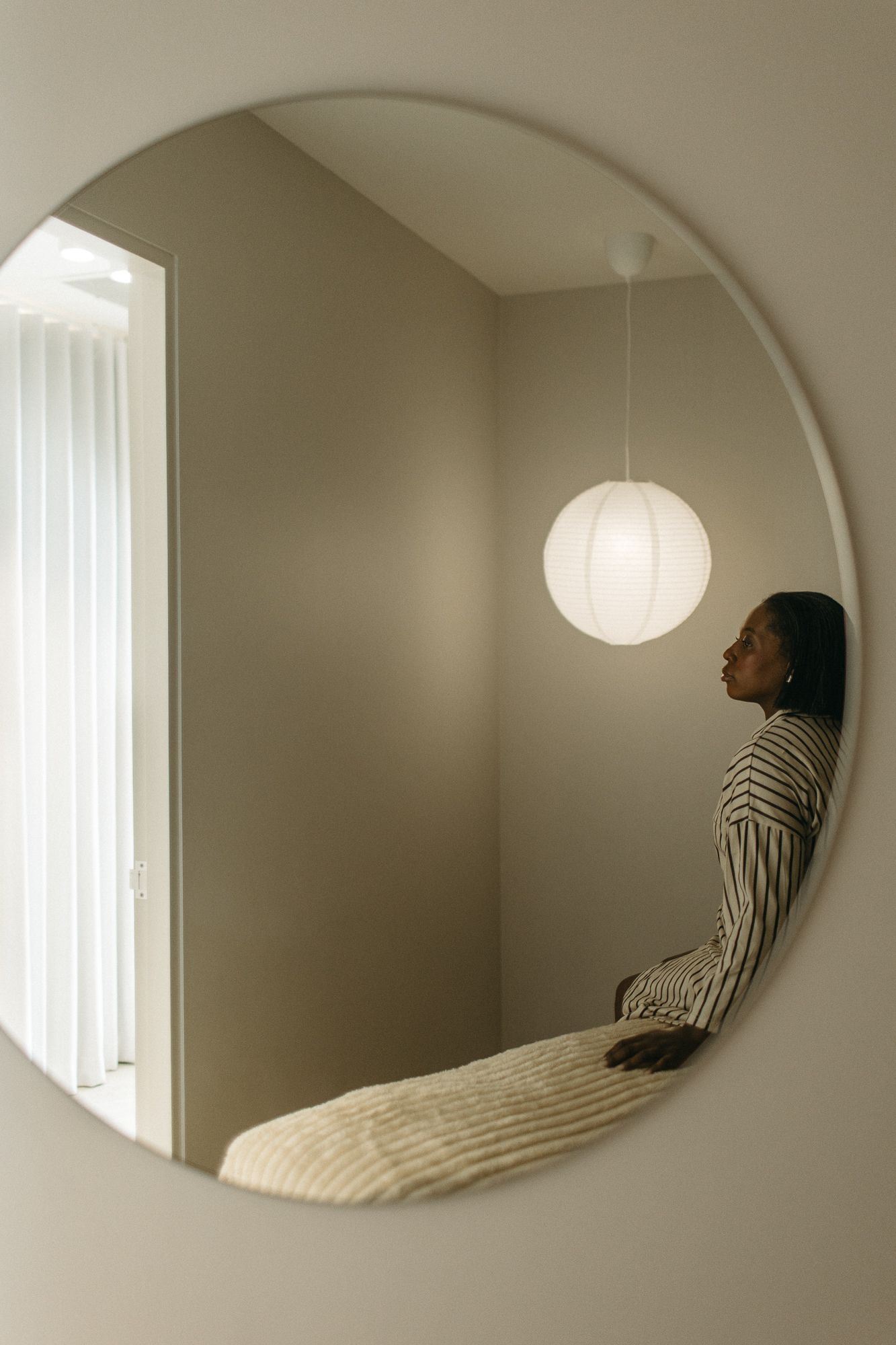 A woman in a striped shirt reflected in a round mirror with a glowing pendant light in a minimalist room.