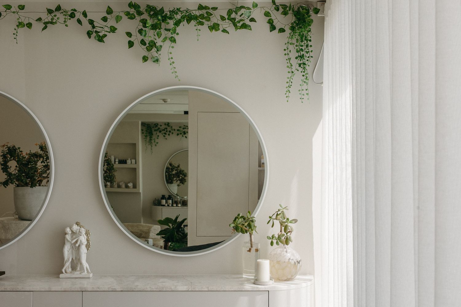 Elegant white bathroom with round mirror, trailing green ivy, marble shelf, and sheer curtains in a bright minimalist space.