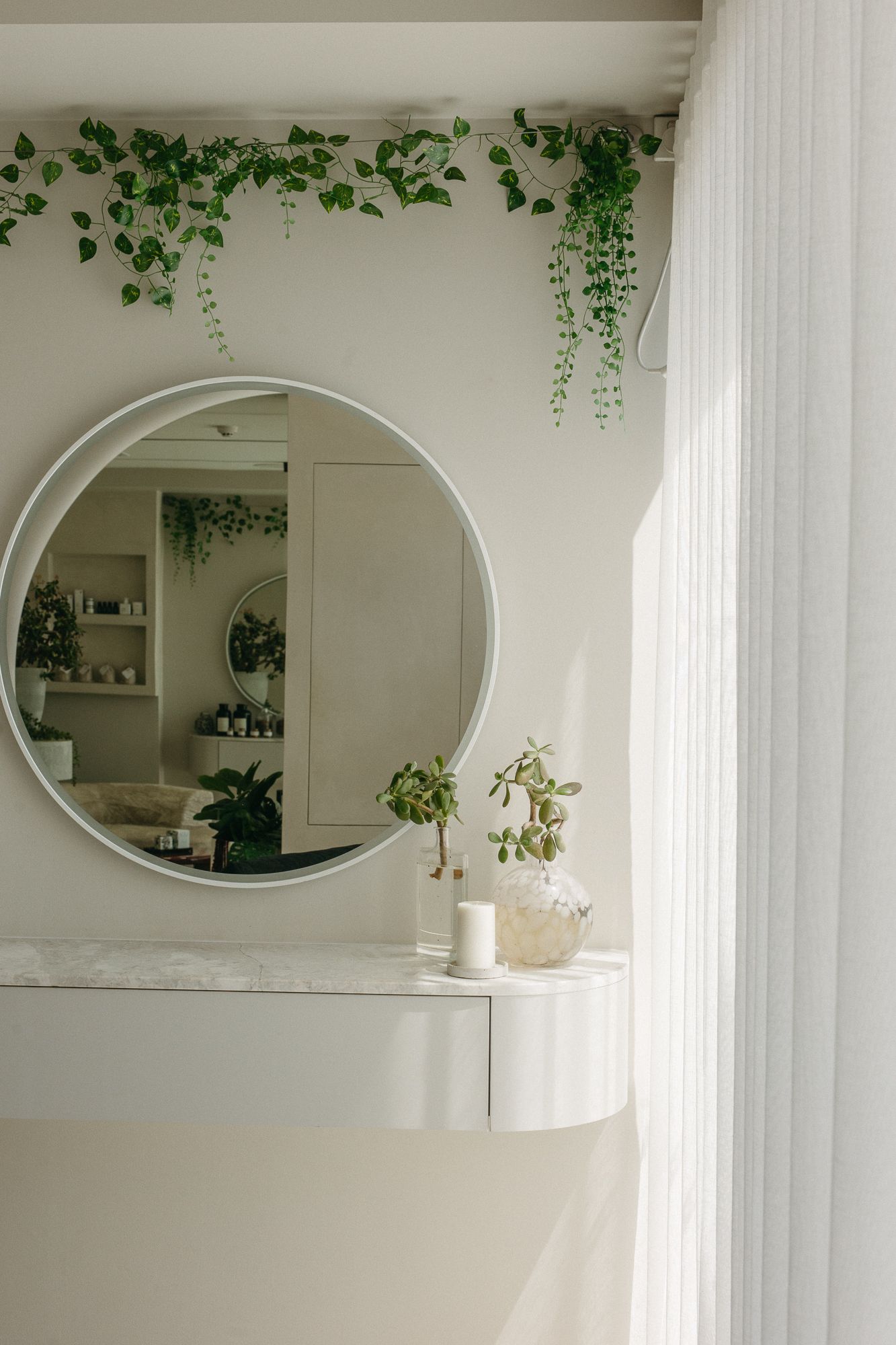Round mirror on white dresser with trailing green ivy, small plant, and candle in bright minimalist room with sheer curtains.