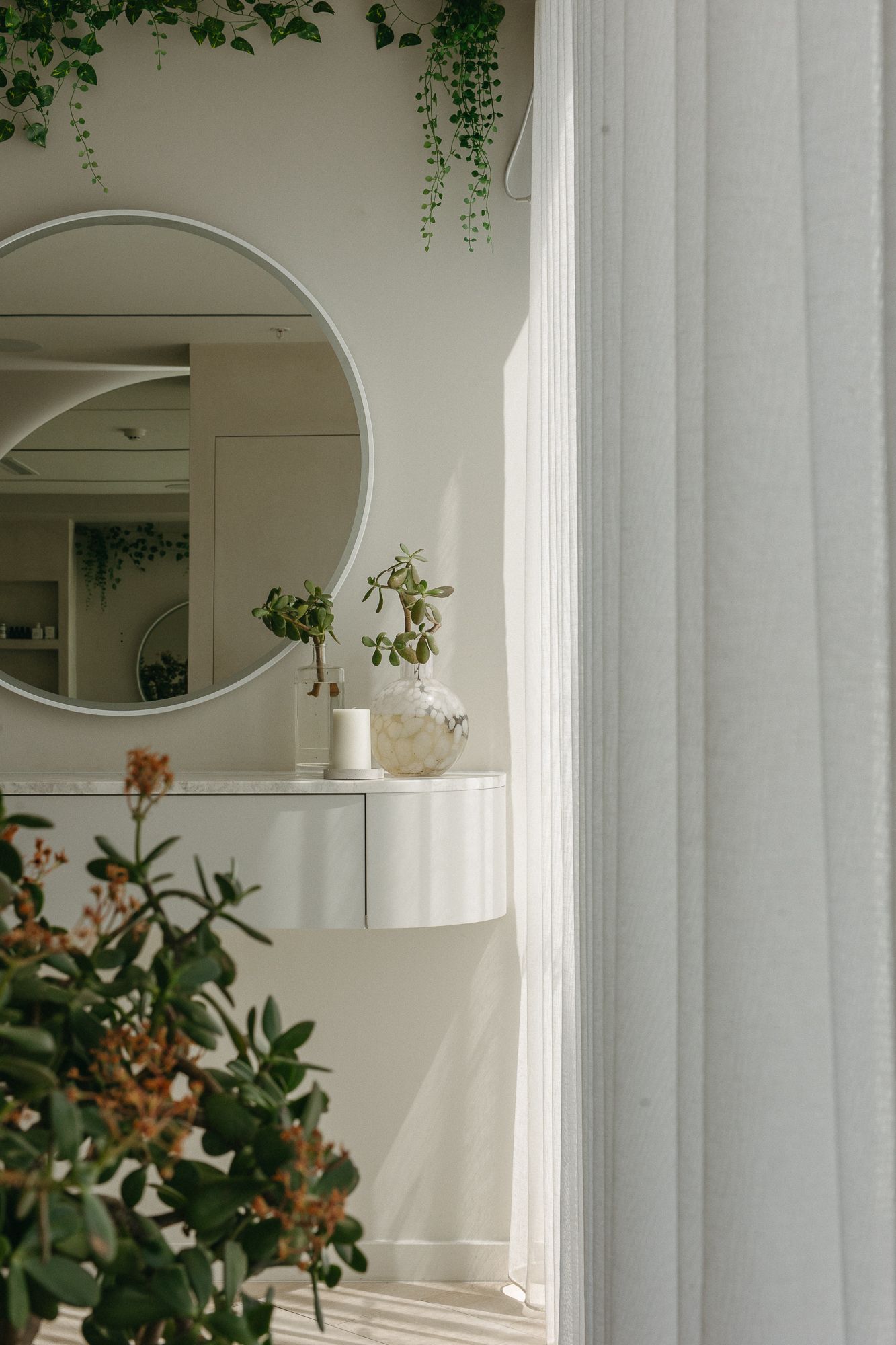 Elegant white bathroom vanity with round mirror, potted plants, and sheer curtains in a bright, minimalist interior.