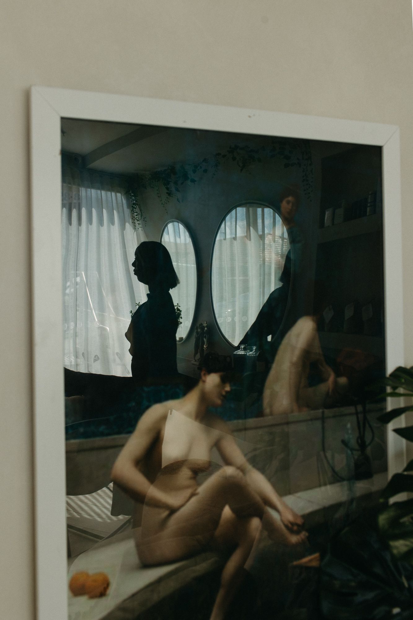 Framed photograph on wall showing a nude figure seated, with silhouettes reflected in an oval mirror in a softly lit room.