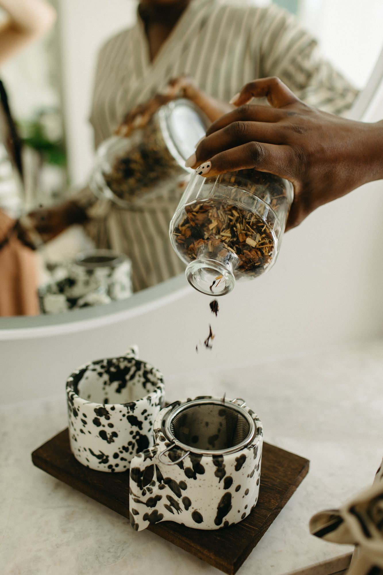 Hands pouring loose leaf tea from a glass jar into a speckled ceramic mug on a wooden tray.