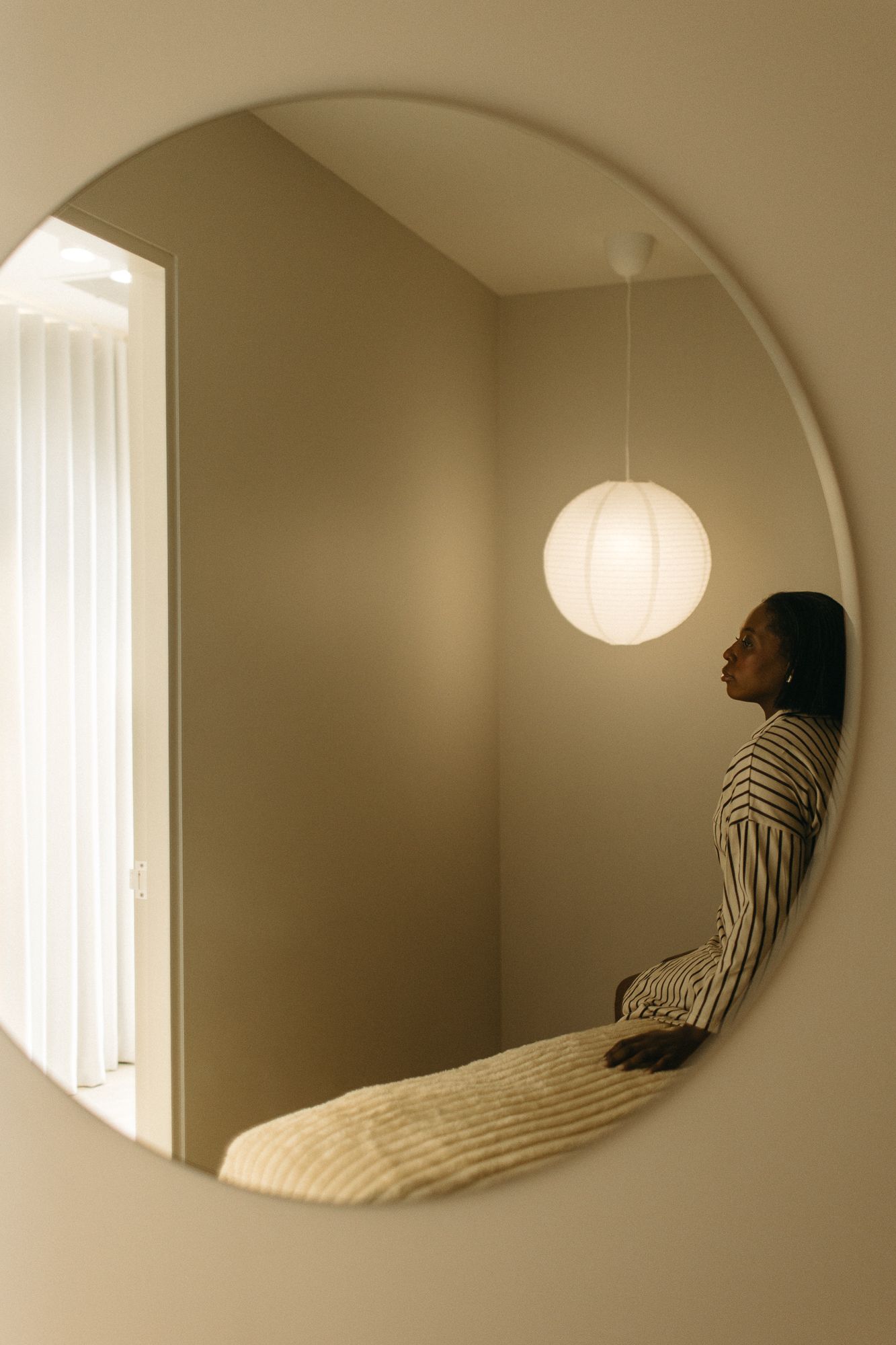 A woman in a striped shirt reflected in a round mirror with a glowing pendant light in a minimalist room.