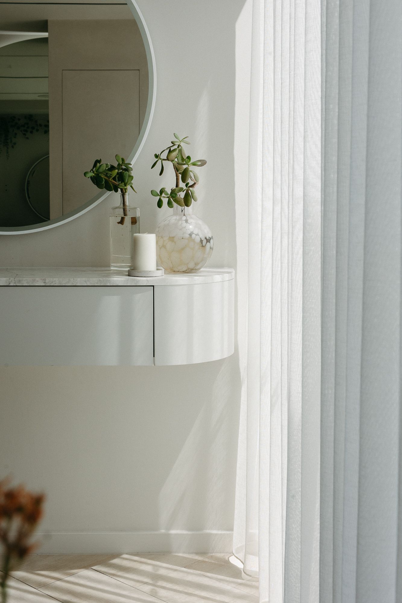White floating vanity with round mirror, candle, and green plants in a bright, minimalist room with sheer curtains.