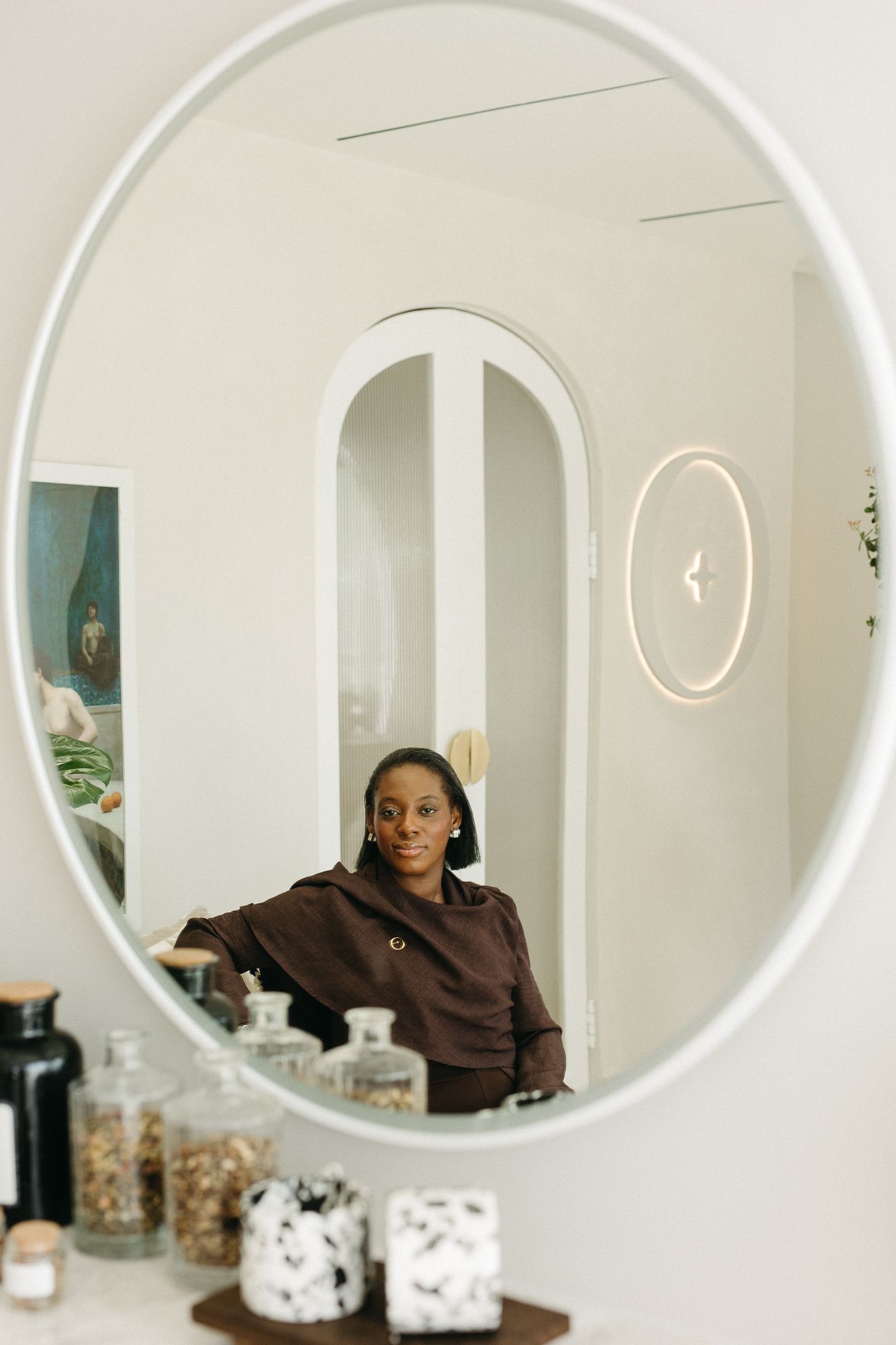 A woman in a brown top is reflected in a round white-framed mirror, surrounded by decorative glass jars.