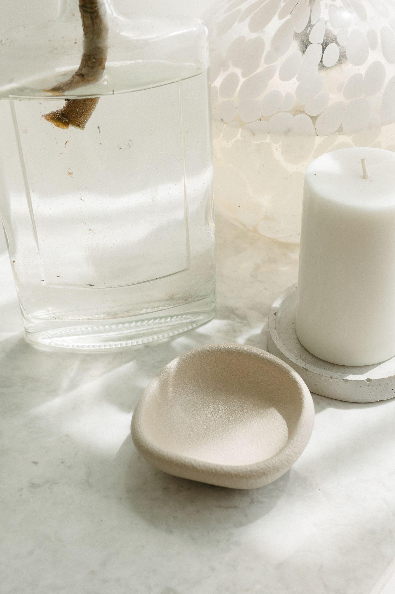 Minimalist spa setup with glass vase, white pillar candle, and beige ceramic dish on marble surface.
