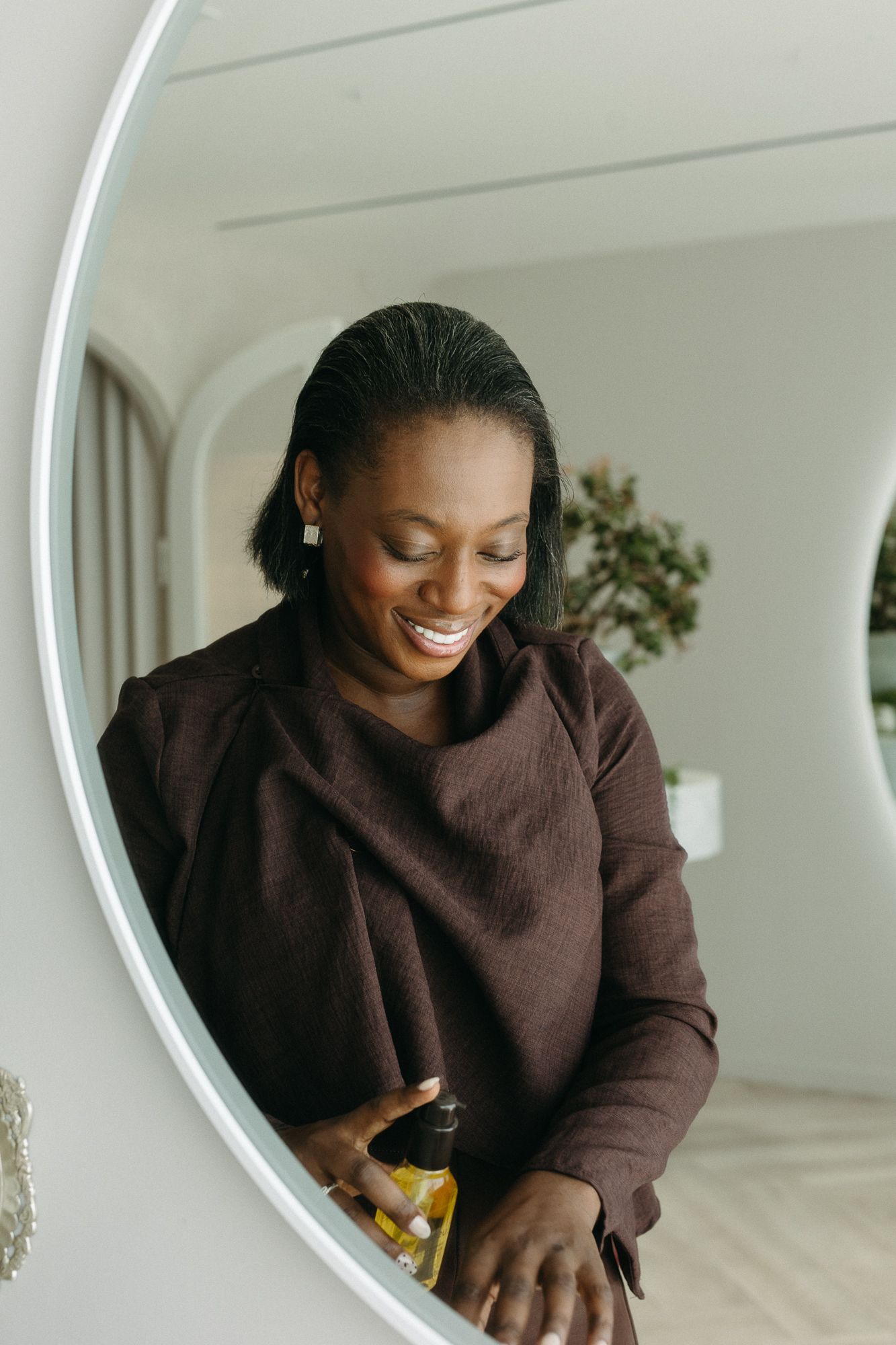 Smiling Black woman in burgundy sweater holding a bottle, reflected in a round LED bathroom mirror.