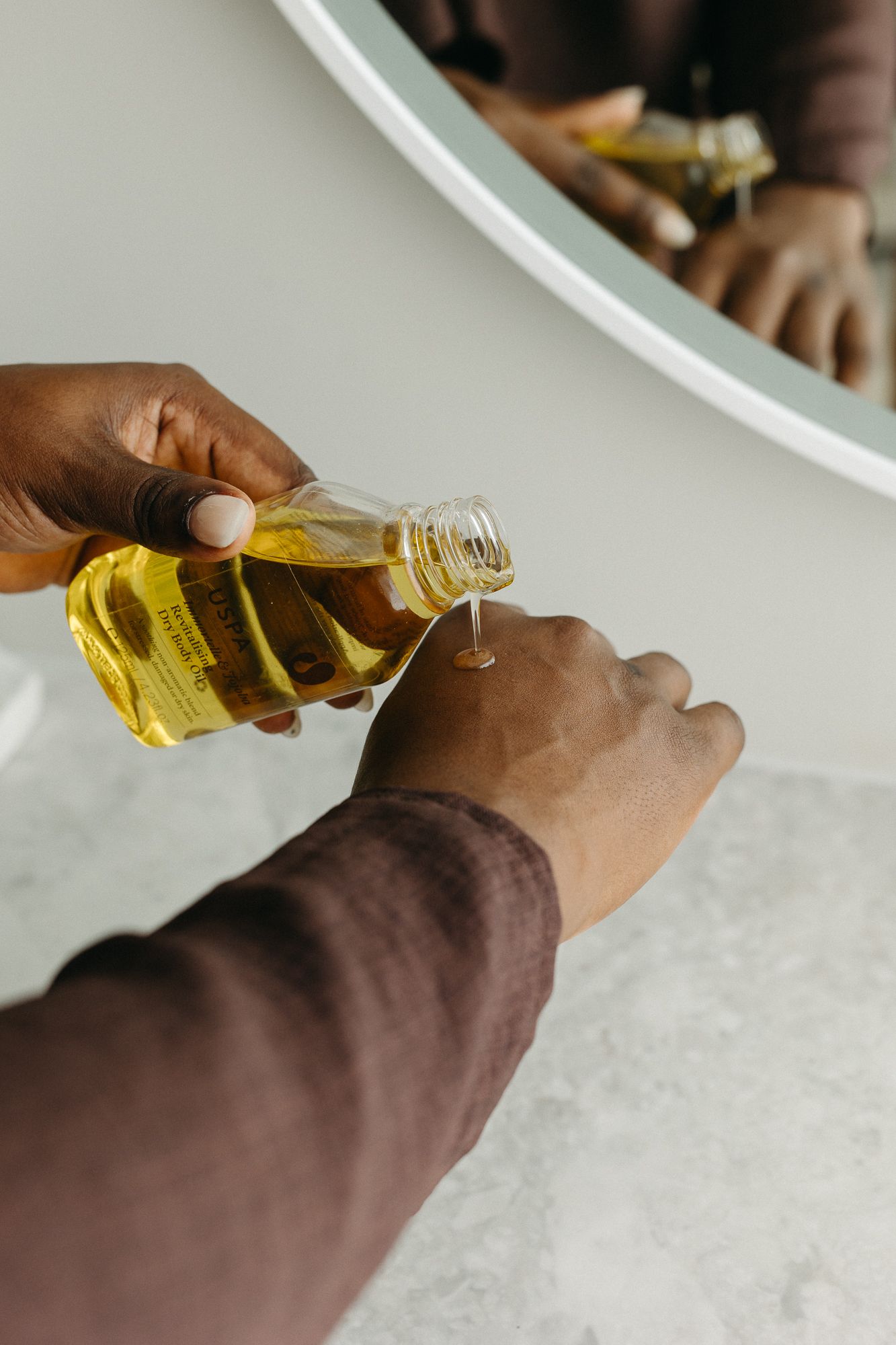 Person pouring golden oil from a small bottle into their hand, reflected in a round bathroom mirror.
