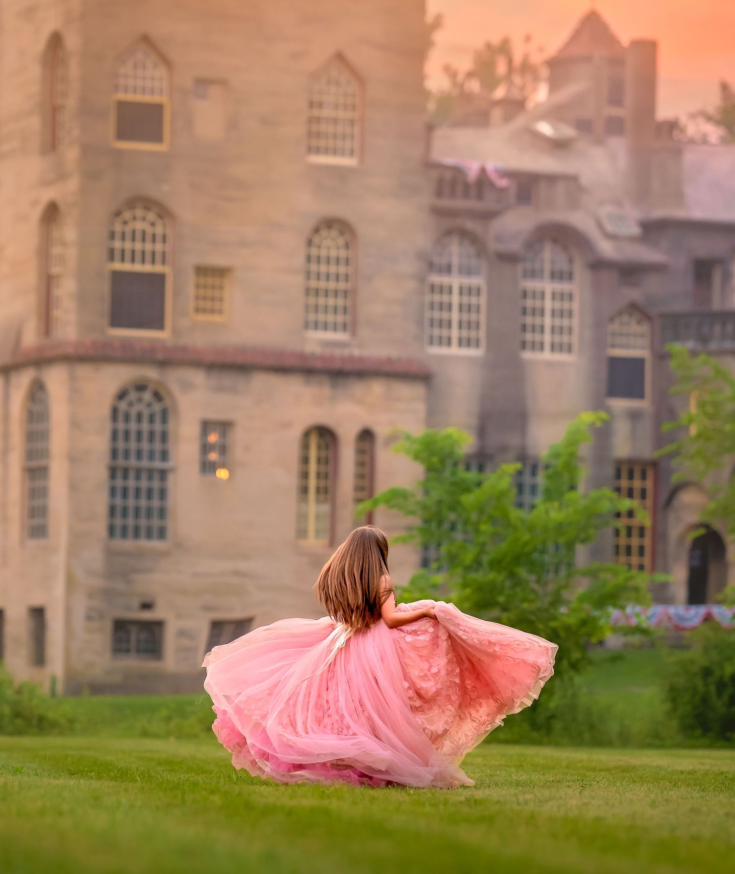 A girl in a flowing pink gown twirls on green grass before a grand castle at sunset.