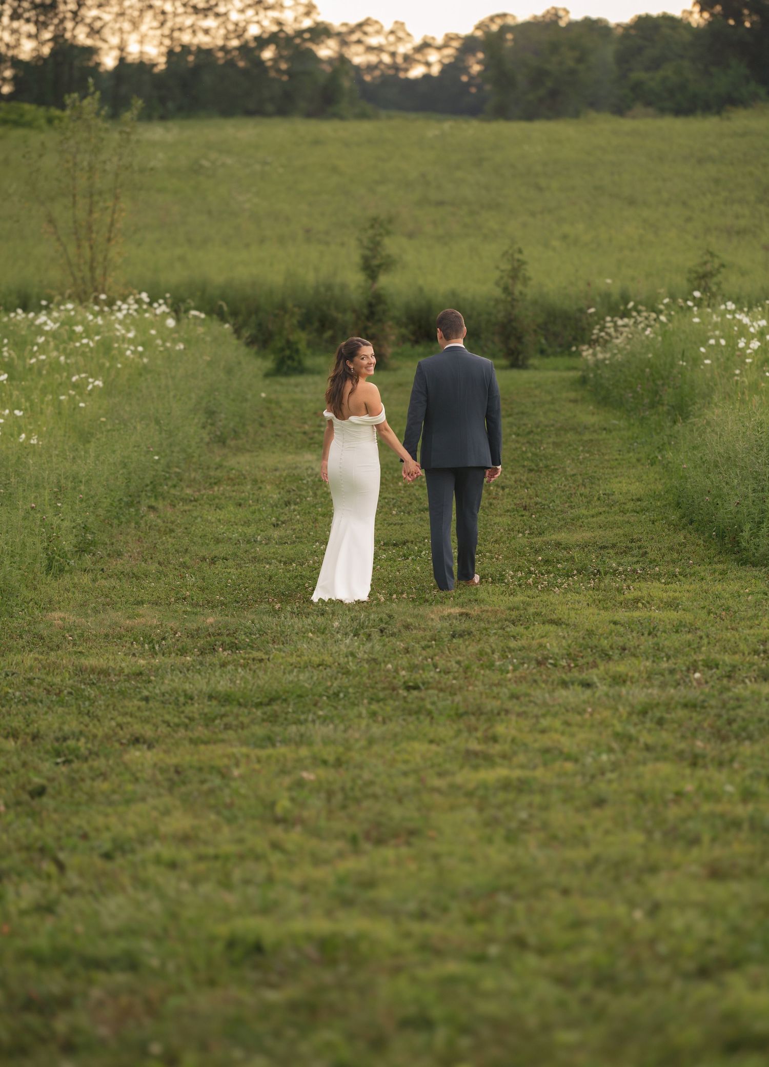A bride and groom walk hand in hand through a lush green field at sunset on their wedding day at farmhouse at Magnolia Hill Farm, Ohio