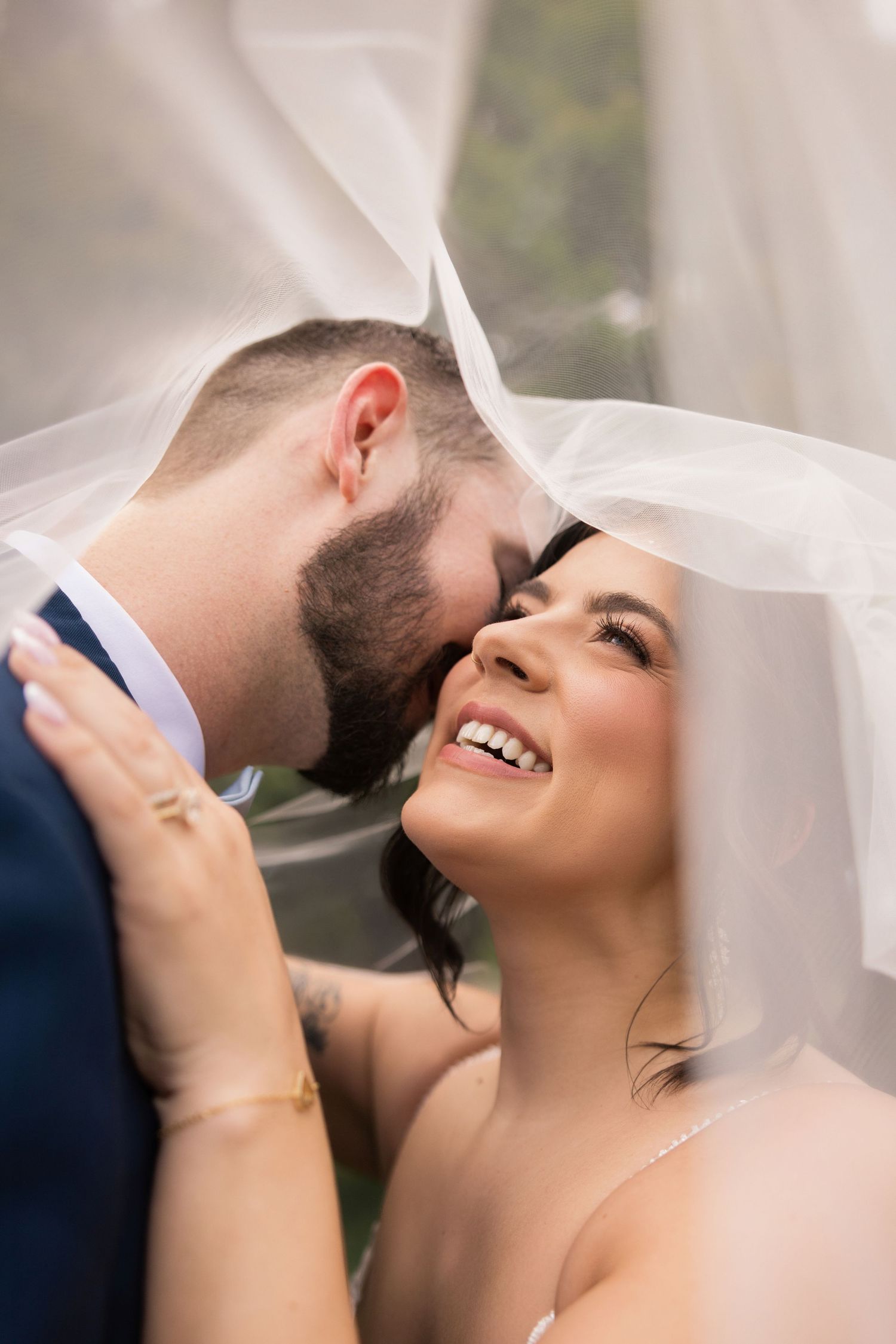 Bride smiling joyfully under veil as groom leans in for a kiss on their wedding day.