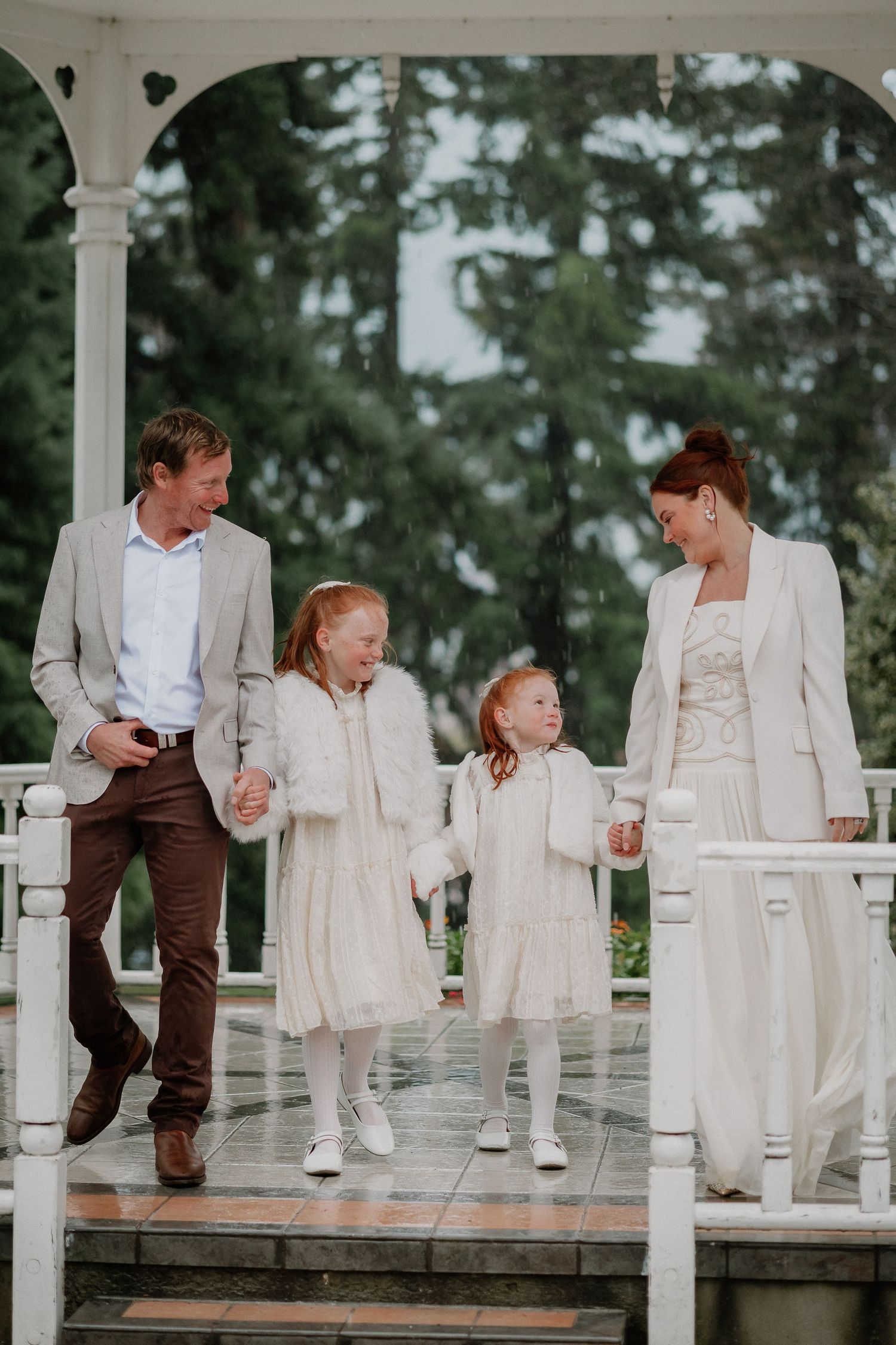 Family of four in cream and neutral outfits holding hands on a white gazebo with evergreen trees behind, a parent and two young girls.