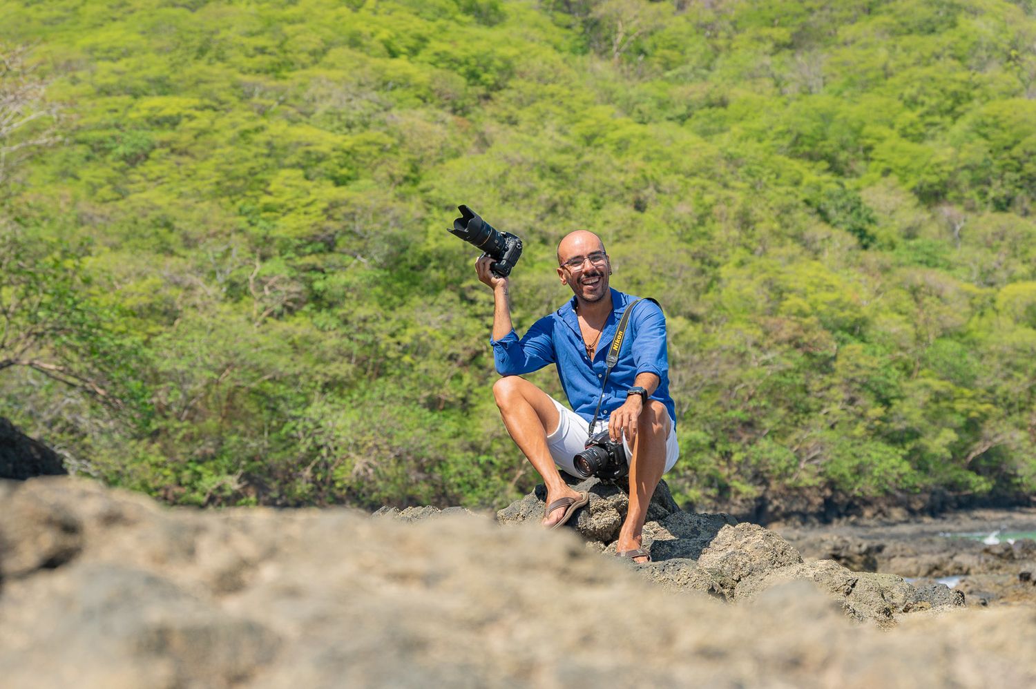 Man crouching on rocky terrain holding camera up, smiling with lush green tropical hillside in background.