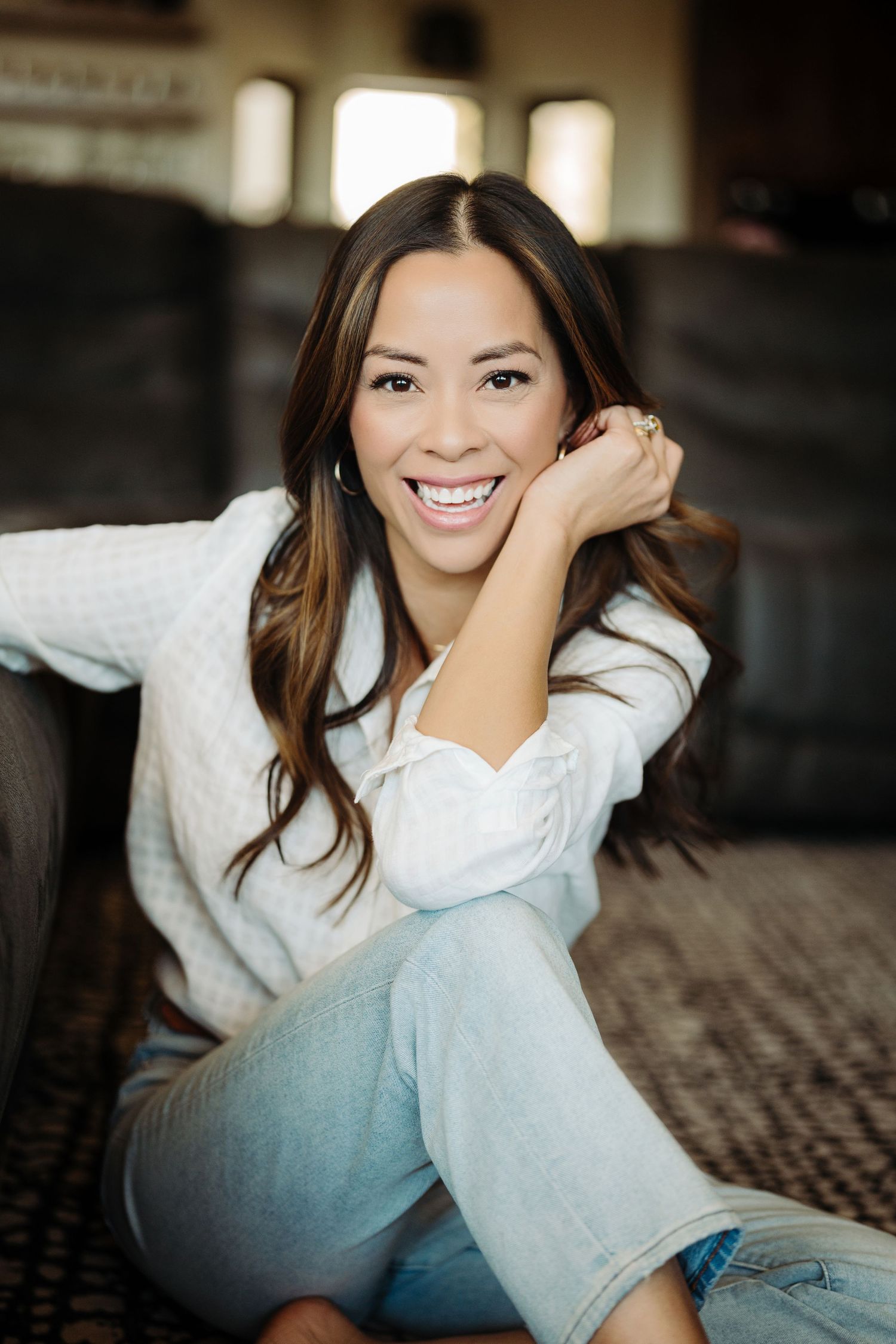 Smiling young woman with long dark hair sits on floor in white sweater and jeans, leaning on couch.