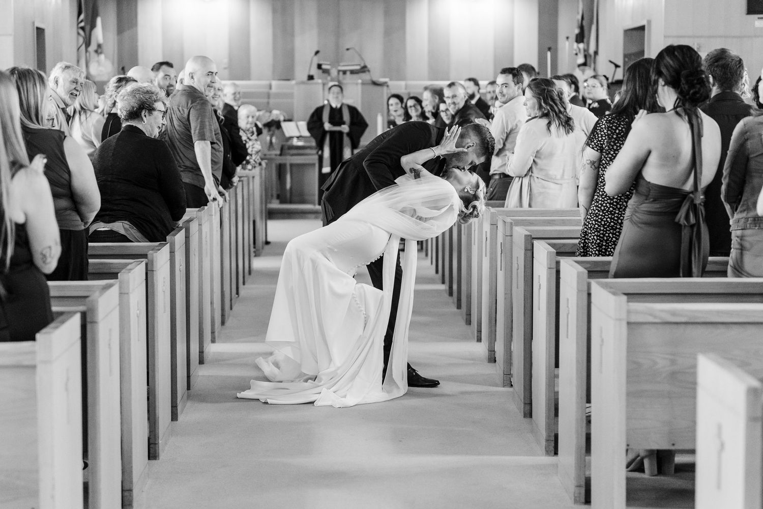 Black and white photo of bride and groom sharing a romantic dip kiss in church aisle during wedding ceremony.